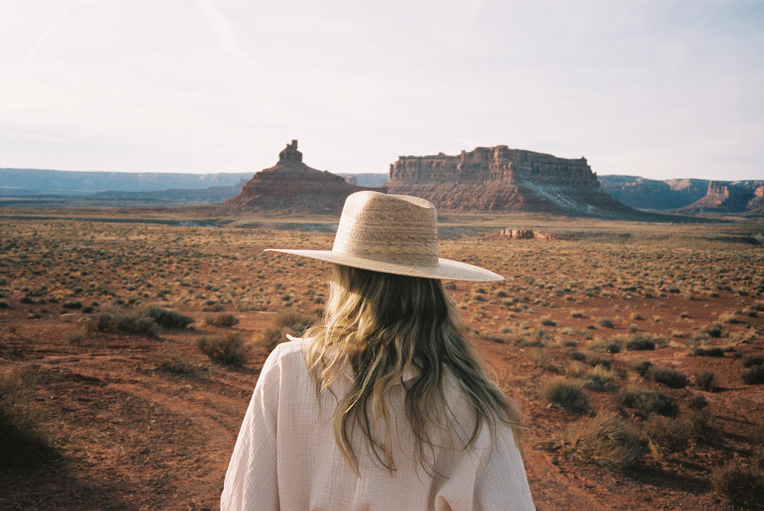 A woman with long blonde hair wearing a wide-brimmed straw hat and a white long-sleeve shirt looking at a desert landscape with mesas and rock formations at sunset.