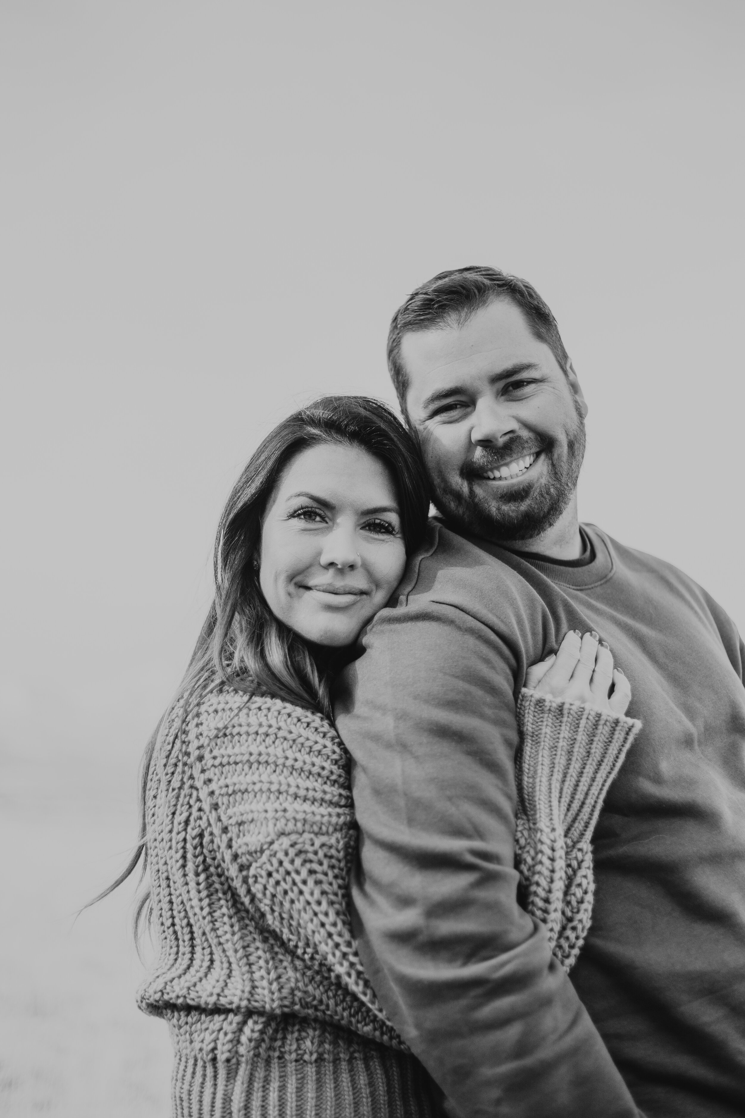 A black and white photo of a smiling couple, the woman with long wavy hair and wearing a knitted sweater, and the man with short hair and a beard, wearing a plain shirt. They are close together, with the woman holding onto the man's arm and shoulders