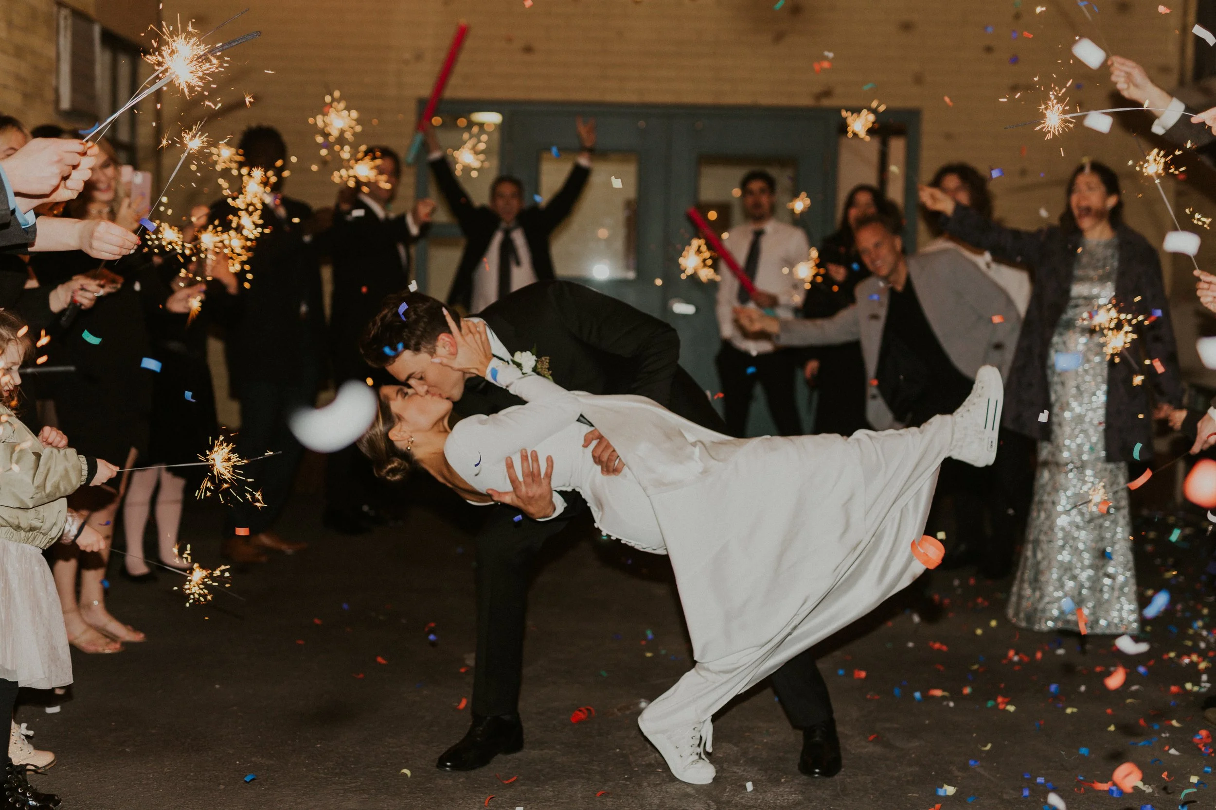 A bride and groom are kissing amid a celebration with sparklers, confetti, and guests cheering in a festive indoor setting.