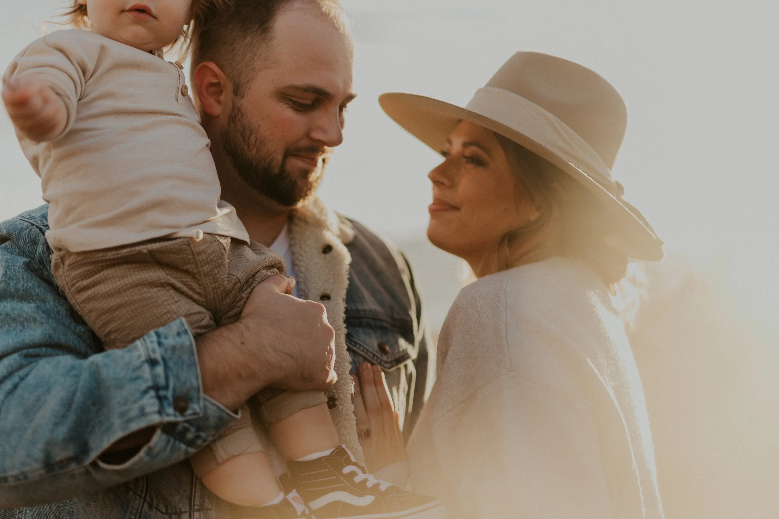 A man holding a young girl on his shoulders while standing outdoors at sunset. The man has a beard and is smiling with his eyes closed, wearing a denim jacket. The girl wears a light-colored hoodie and looks off to the side. The background is bright 