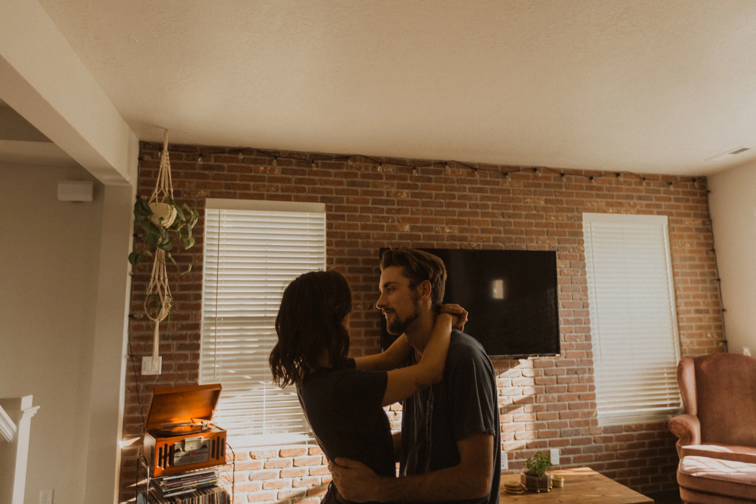 A couple dancing closely inside a living room with brick walls, blinds, and a vintage record player.