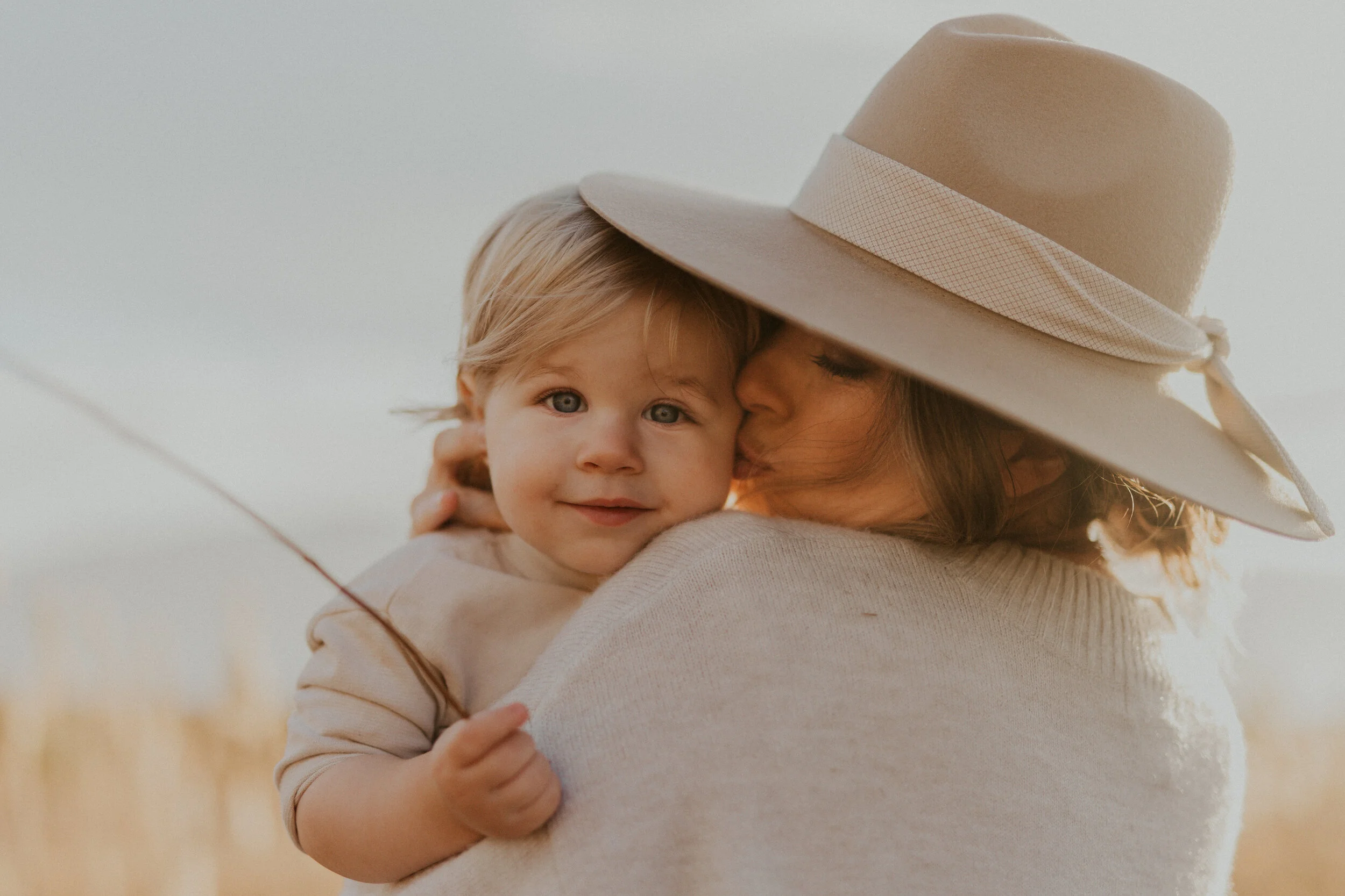 A woman wearing a beige sunhat holding a young child with blonde hair and blue eyes outdoors.