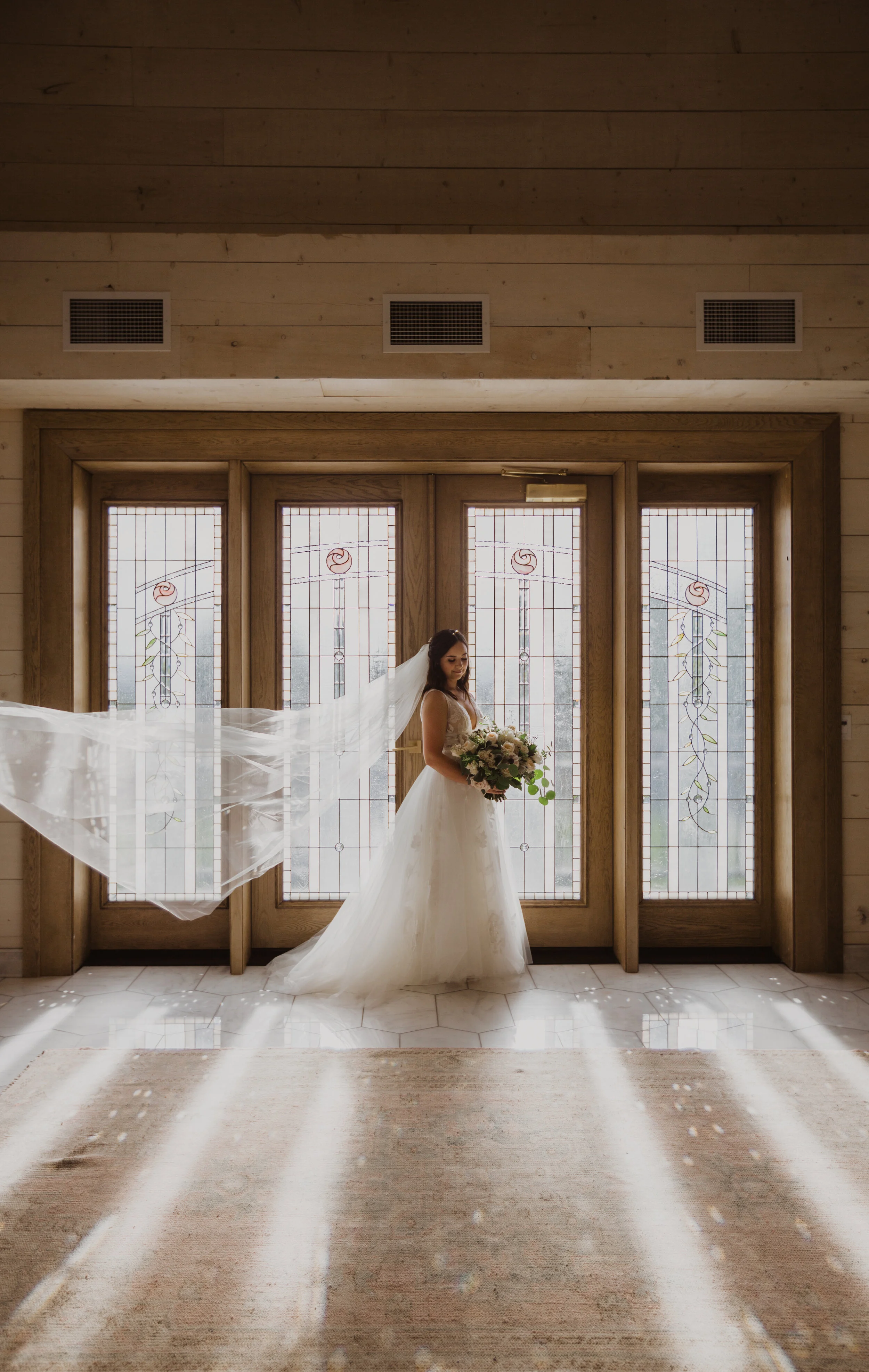 A bride in a white wedding dress holding a bouquet of flowers standing in front of tall stained glass windows in a wooden interior with sunlight streaming in.