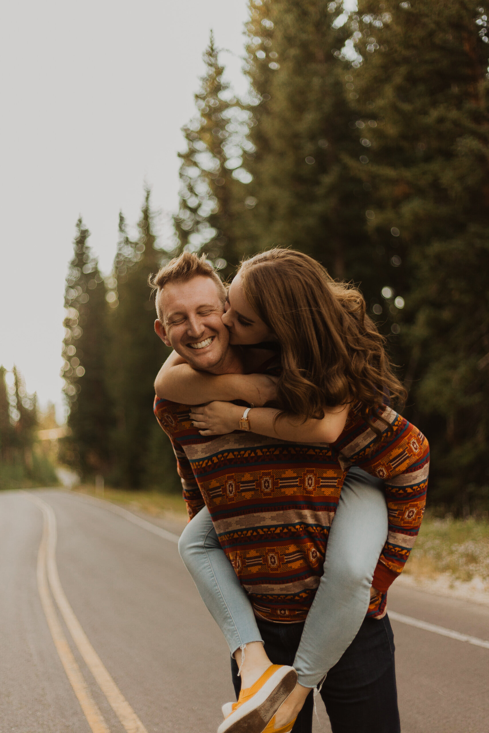 A couple enjoying a piggyback ride on a rural road with tall trees in the background, during what appears to be autumn.
