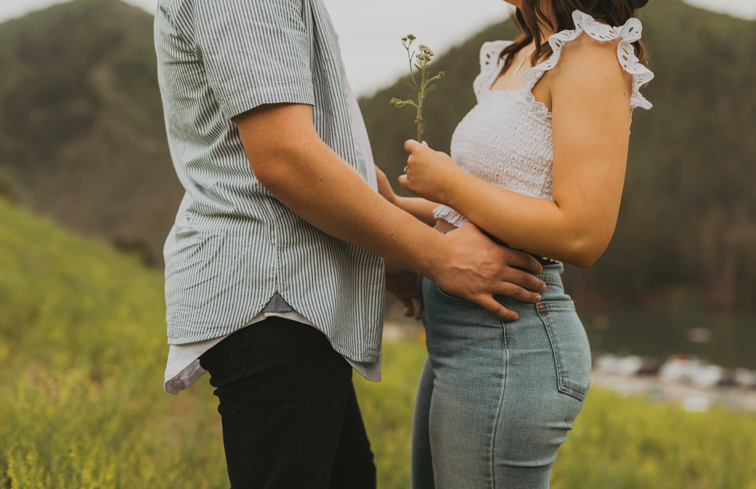 A man and woman holding hands outdoors in a natural setting with grass and trees, the woman is holding small flowers.