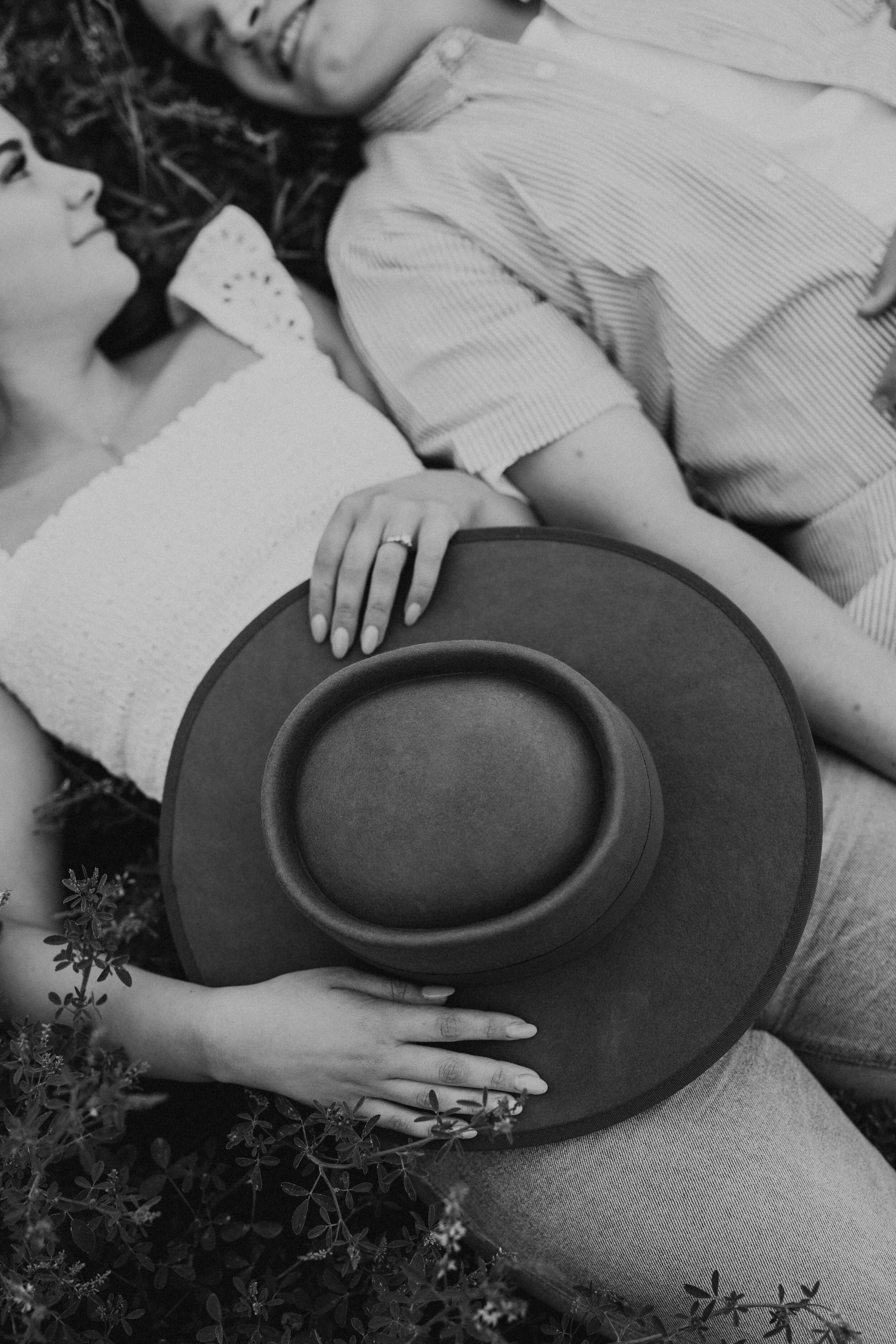 A woman lying on the ground holding a large round hat with a wide brim, with her hand resting on her stomach and her head turned toward a man, who is lying next to her on the grass covered with plants. They are facing each other in an outdoor setting