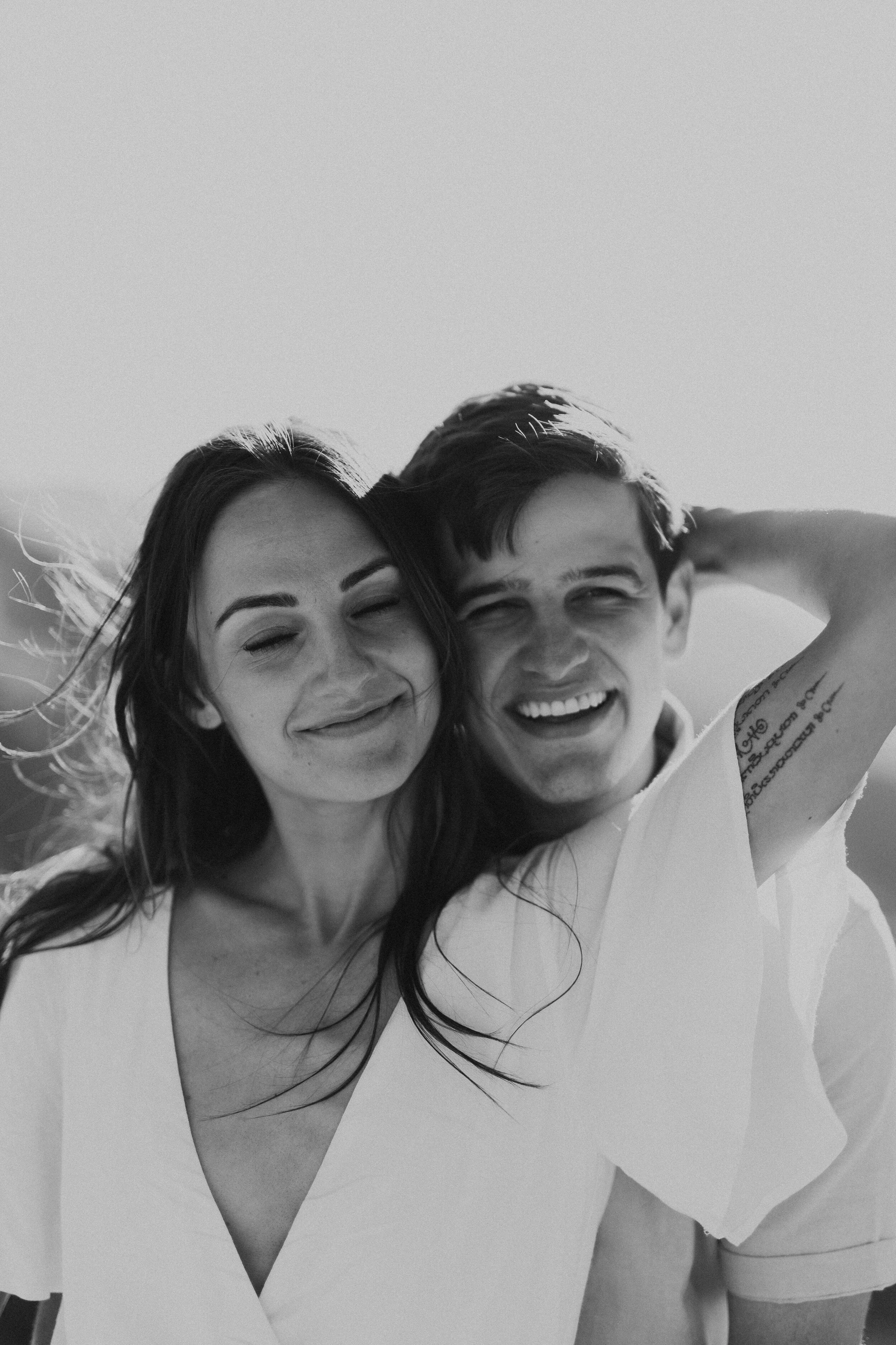 Black and white photo of a smiling woman and a smiling man, both with their eyes closed, leaning close together, outdoors in natural light.
