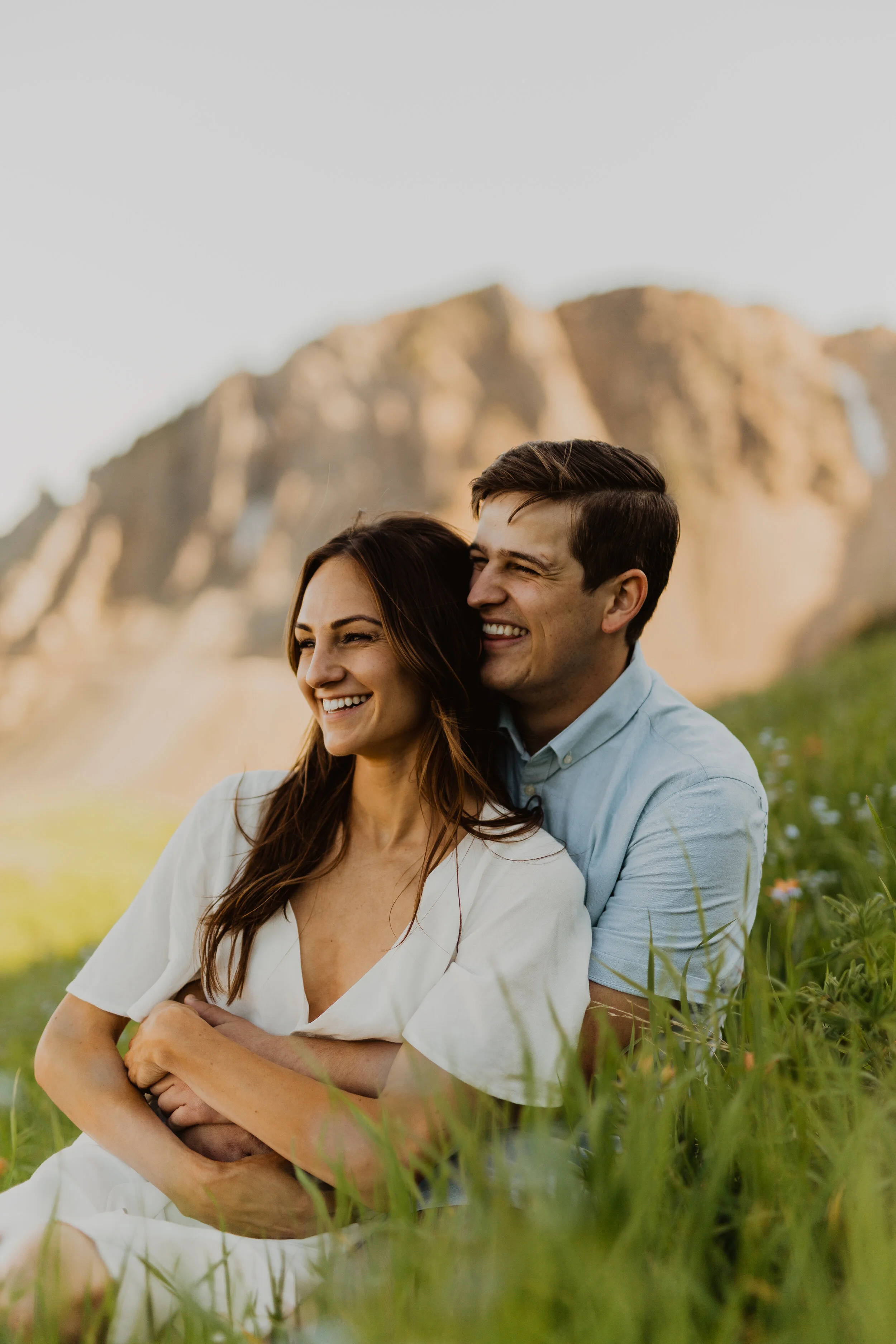 A smiling couple sitting in a grassy field with mountains in the background, embracing each other.