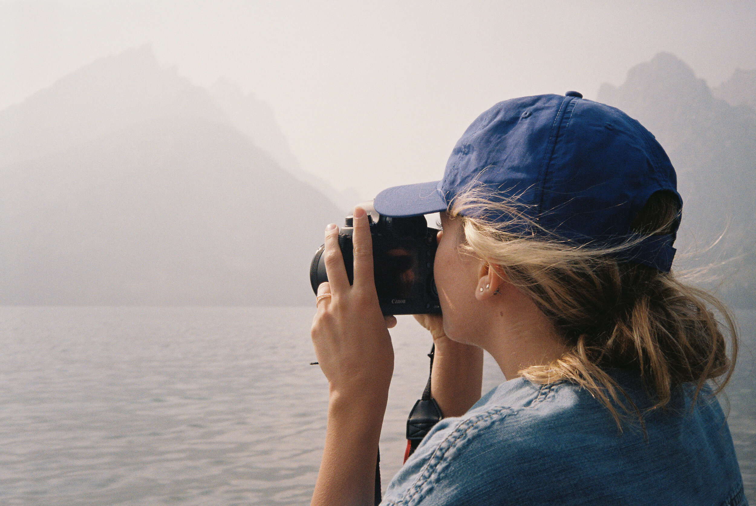 A woman taking a photograph outdoors near a body of water with mountains in the background, wearing a blue cap and denim jacket.