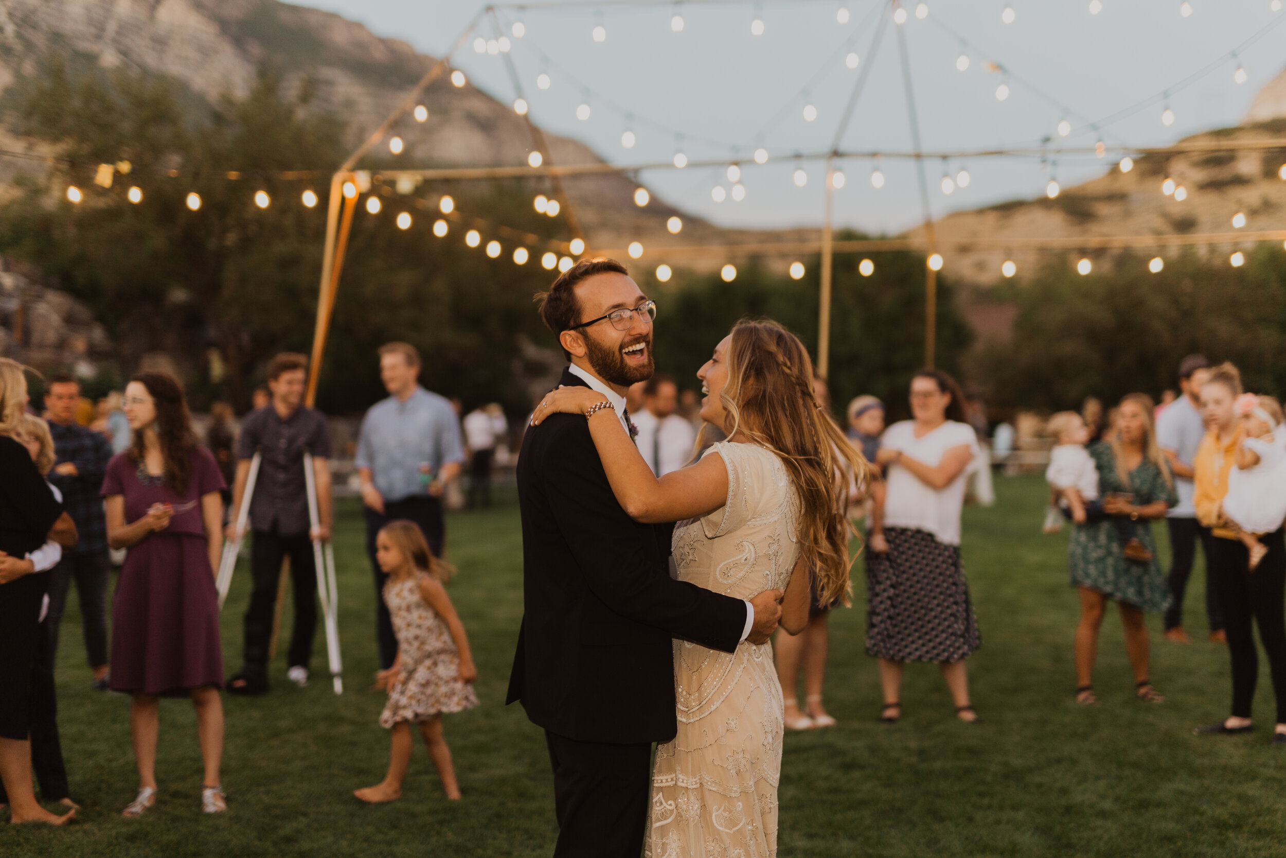 A happy couple dancing together outdoors at a wedding reception at sunset, with string lights hanging overhead and guests in the background.