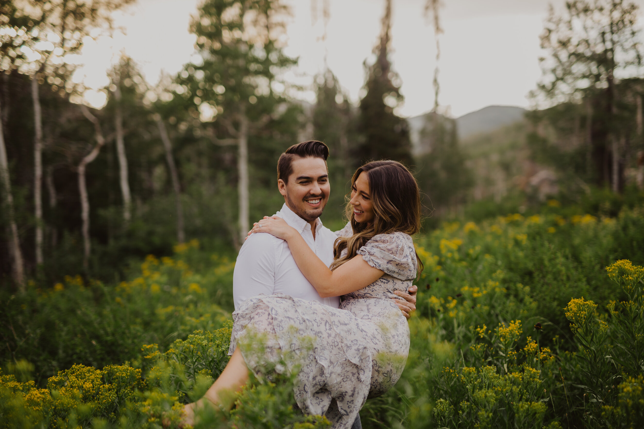 A man and woman embrace in a field of yellow flowers surrounded by trees with sunlight in the background.