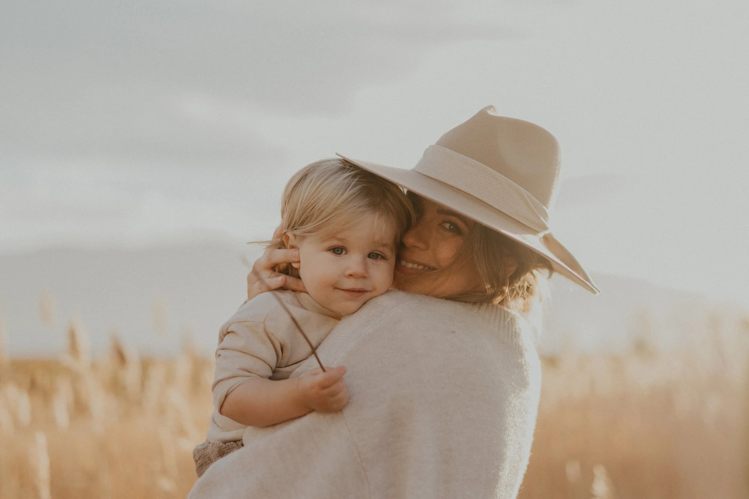 A woman with a wide-brimmed hat holding a young child in a wheat field on a cloudy day.