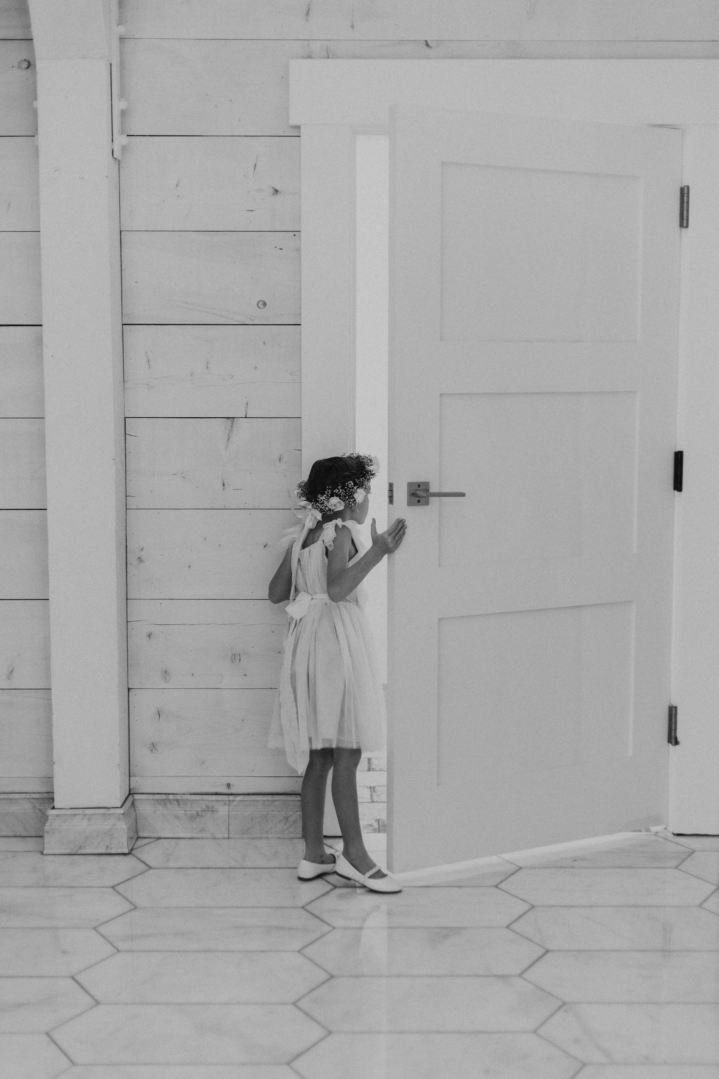 A young girl in a light dress, wearing white shoes, stands next to a large plain white door, with her head down and hands touching the door handle, in a room with hexagonal tiles on the floor and wooden wall panels.