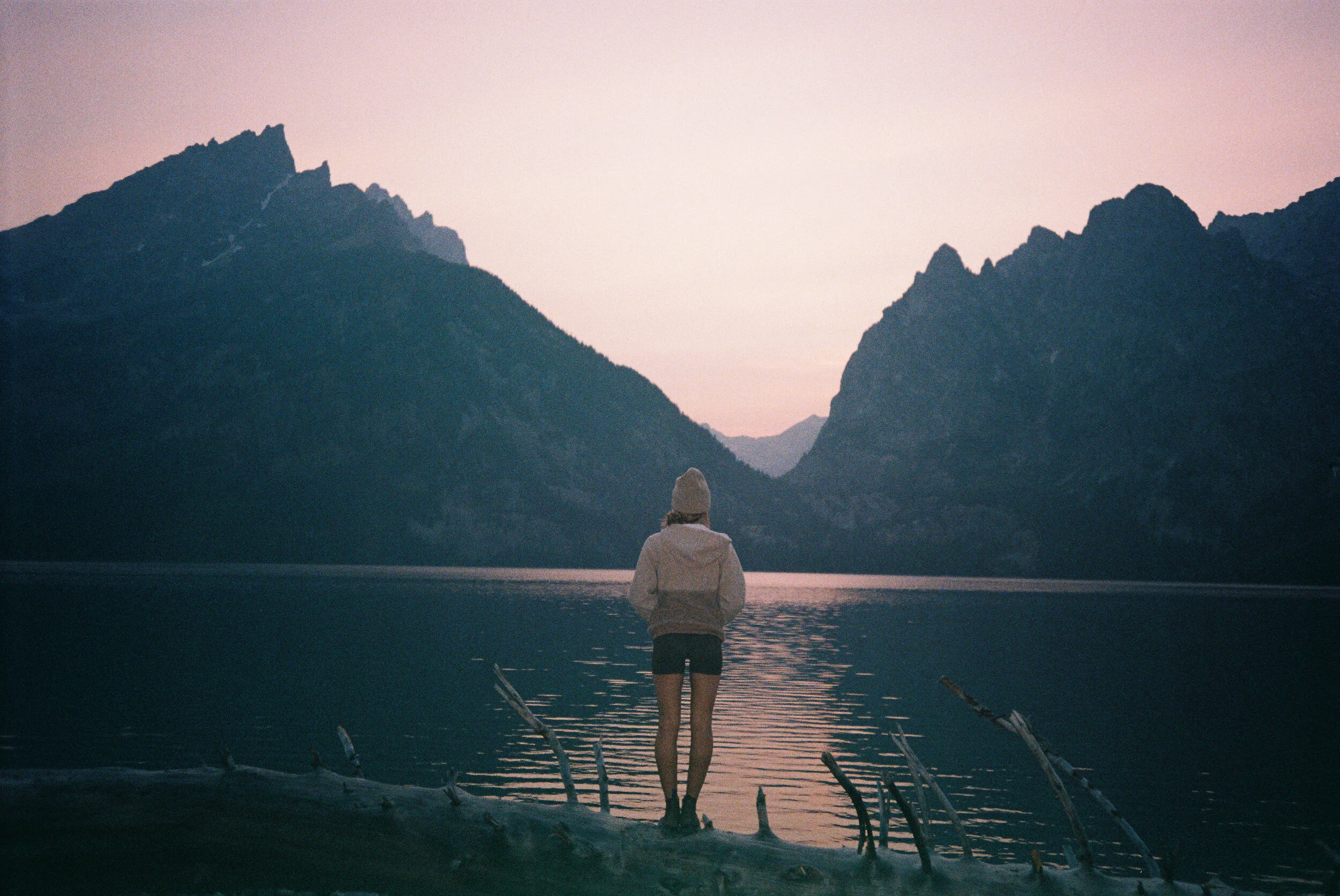 Person standing by a lake at sunset, surrounded by mountains and fallen tree branches.
