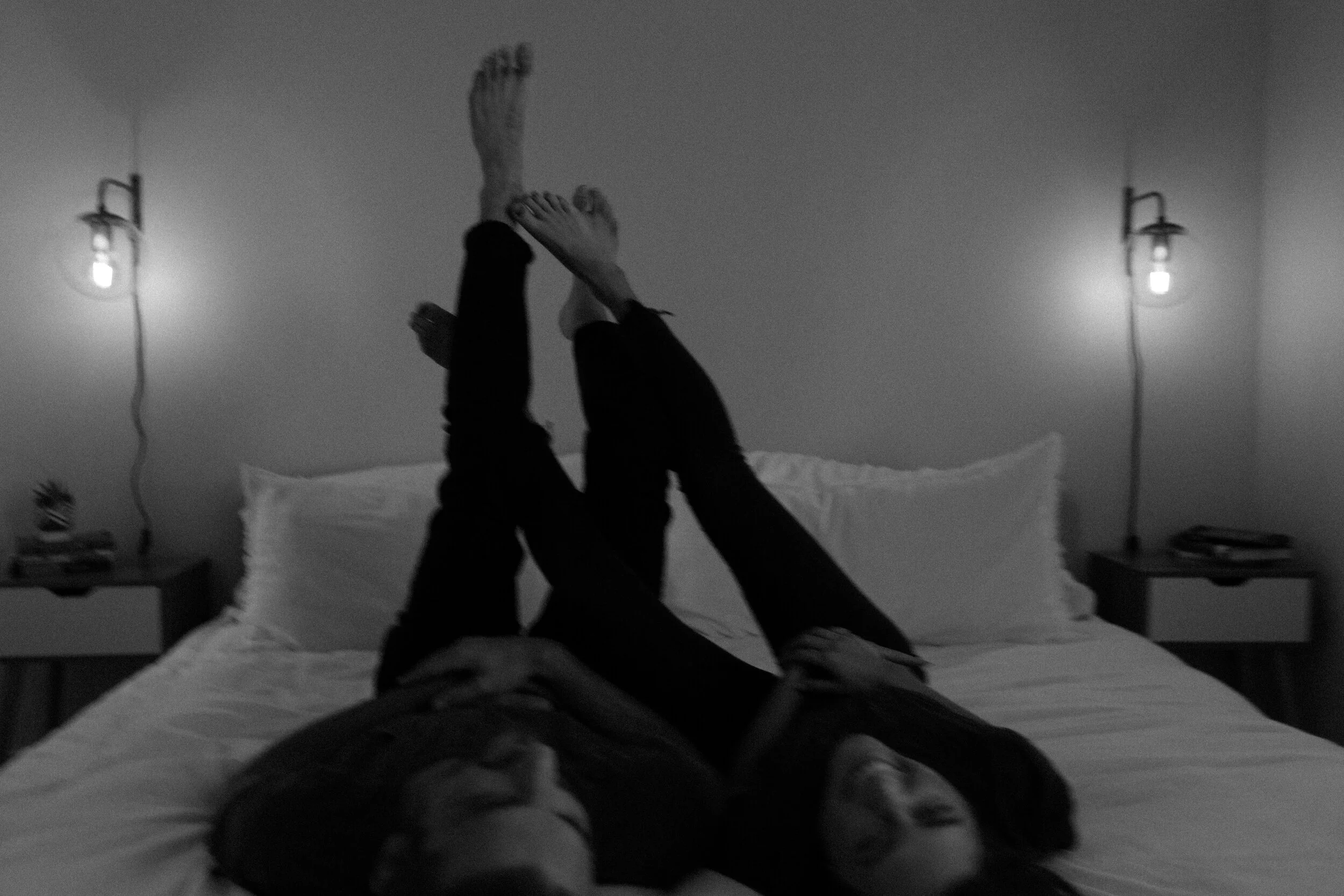 A person lying on a bed with legs raised and crossed at the ankles, wearing dark pants and socks, in a bedroom with two bedside tables and wall-mounted lamps, captured in black and white.