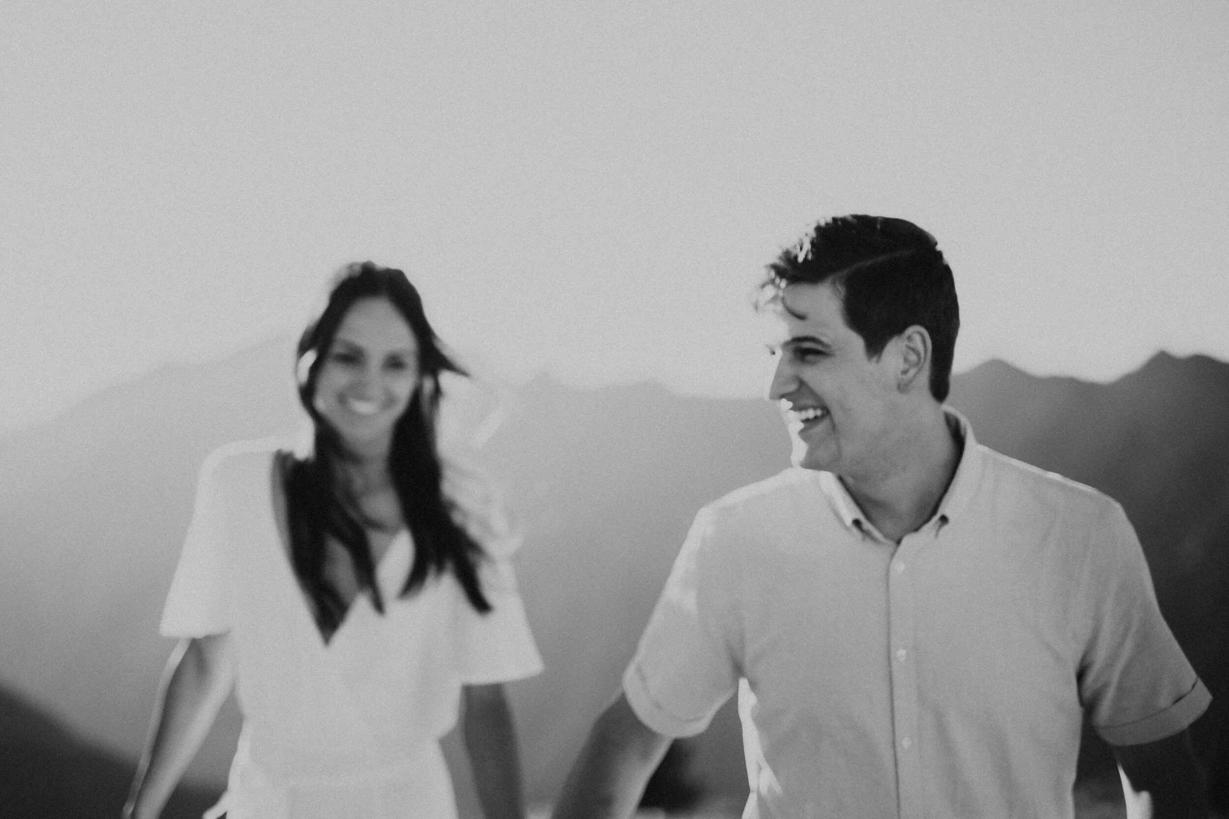 Black and white photo of a man and woman smiling and walking outdoors, mountains in the background.