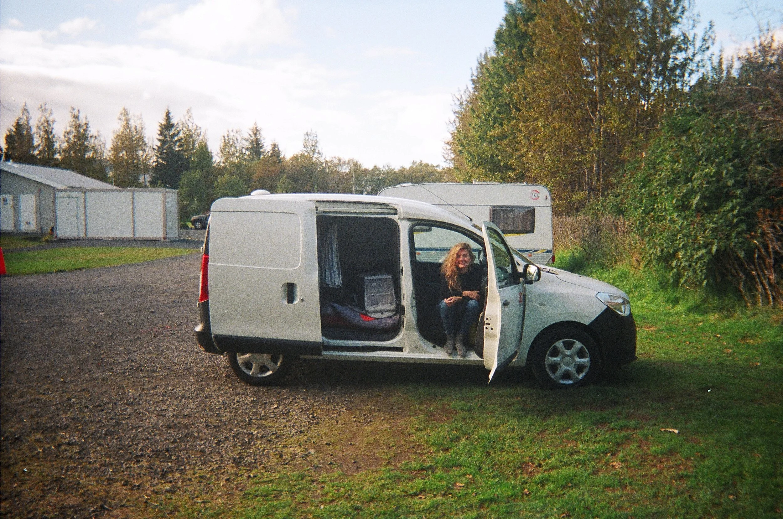 A woman with long curly hair sitting inside a white van with the door open, parked on a grassy area with trees and a recreational vehicle in the background.