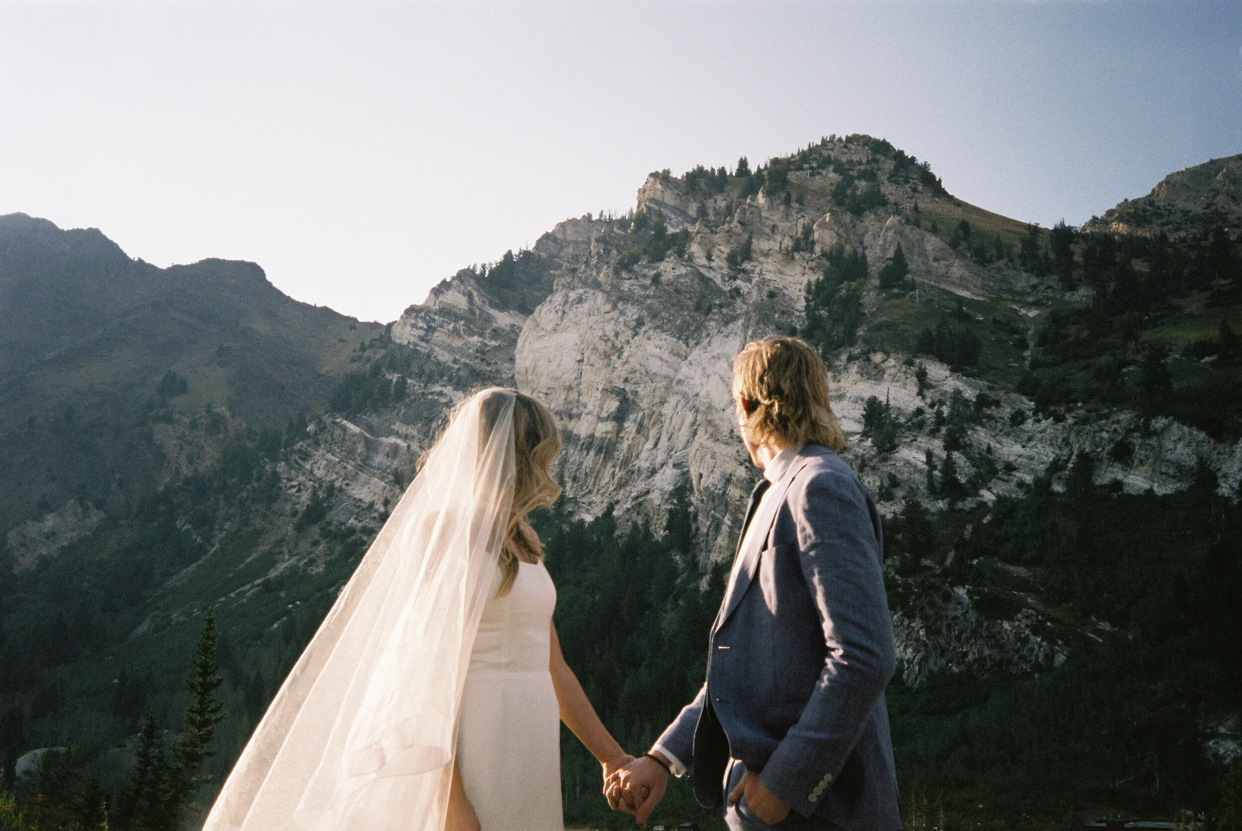 A bride and groom holding hands outdoors against a mountainous landscape.