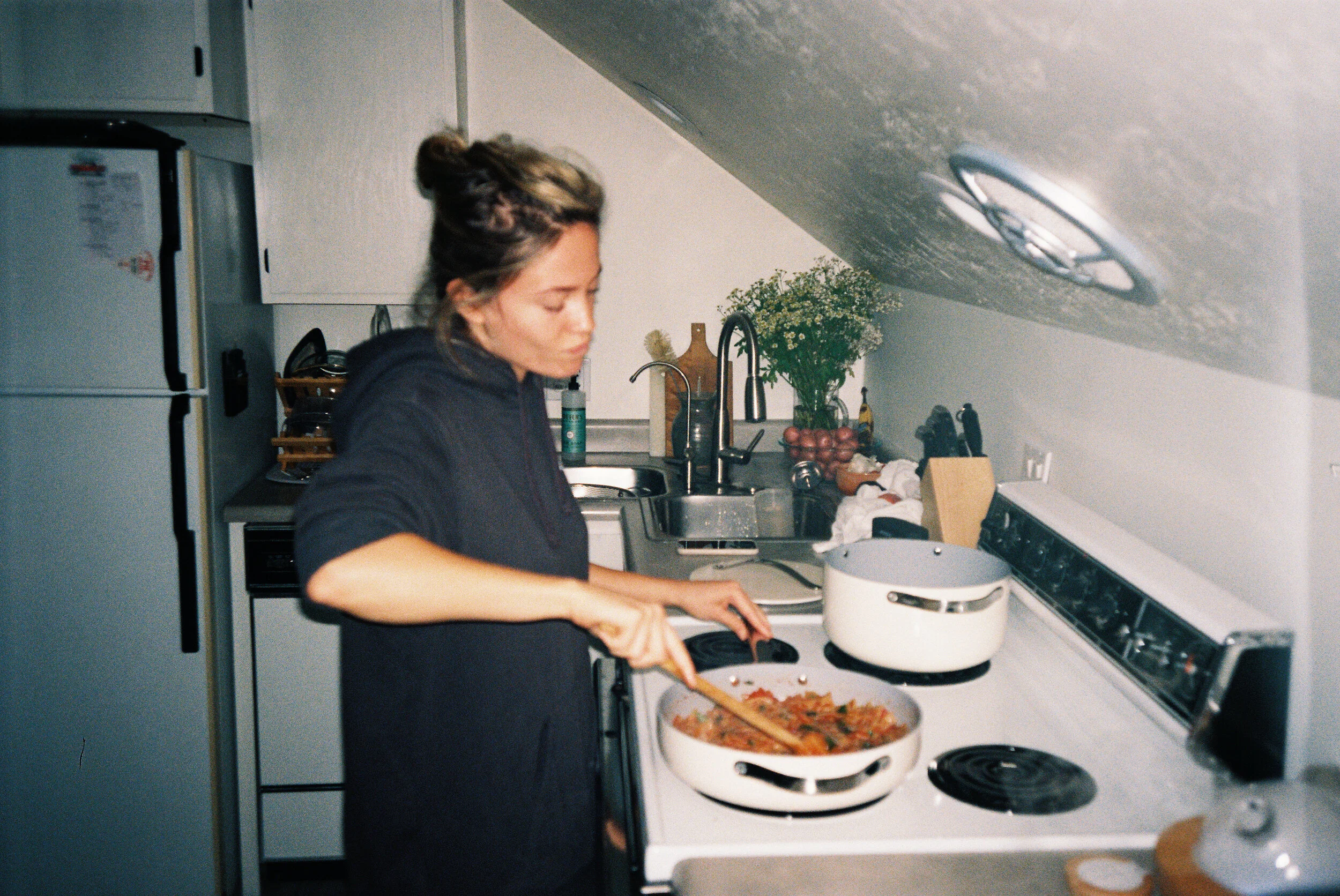 A woman cooking on a stovetop in a kitchen, with a pot of sauce or stew and a pan with food, surrounded by various kitchen items, plants, and decorations.