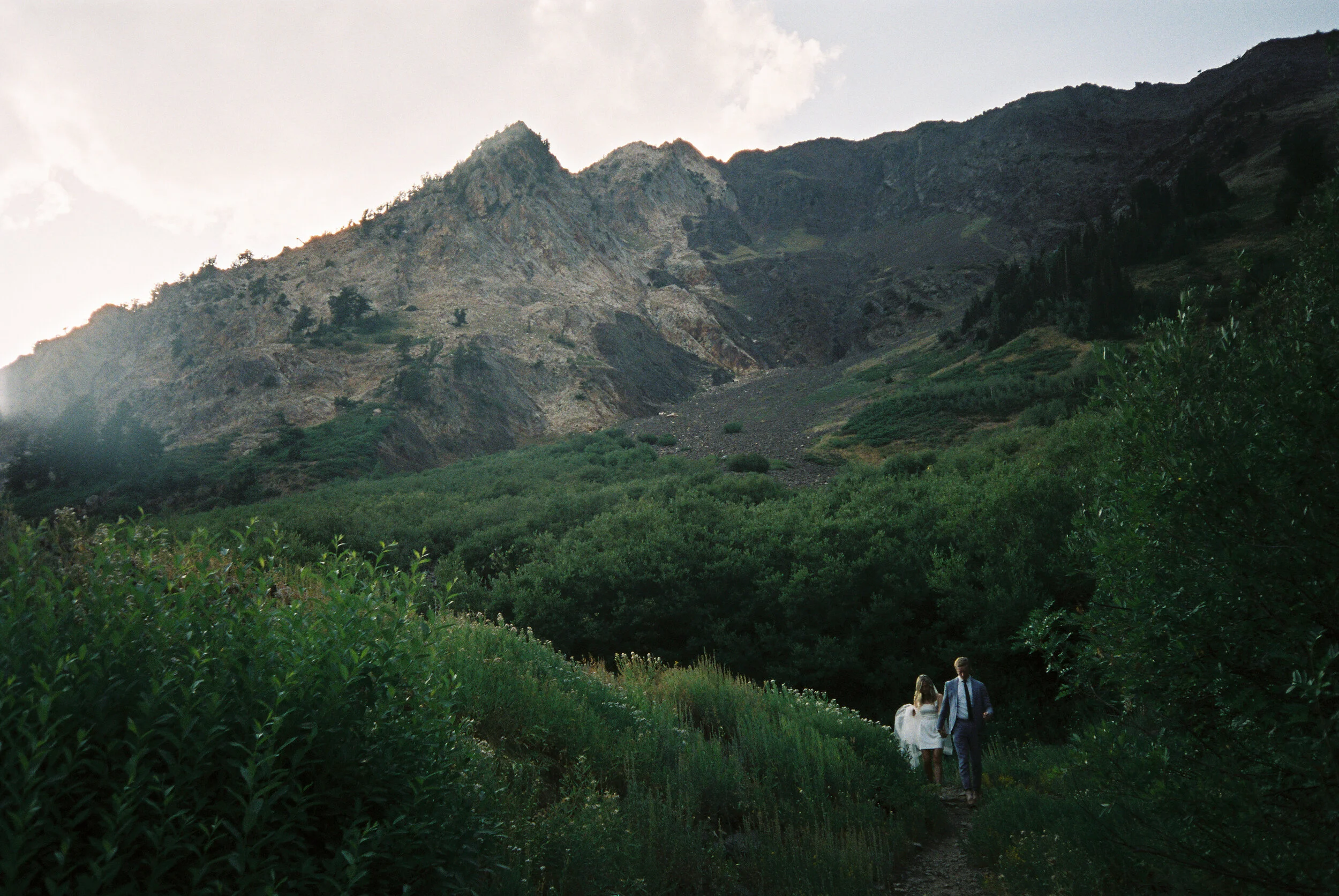 A couple walking on a trail through lush green vegetation with mountains in the background.