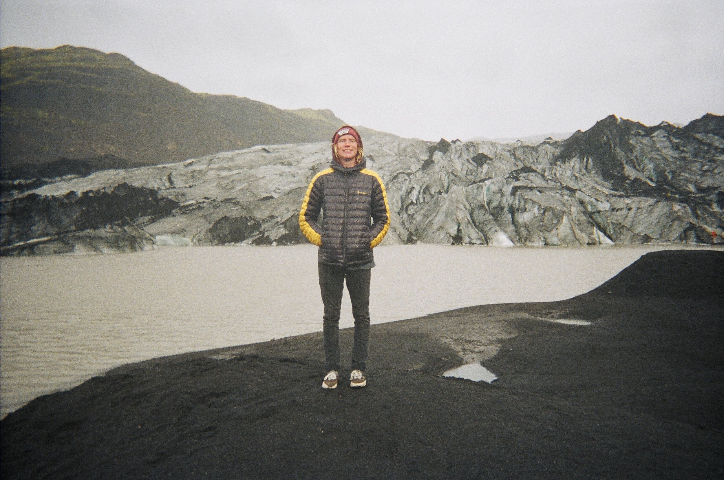A person standing on black volcanic sand, wearing a black and yellow down jacket and a red beanie, with glaciers and rugged mountains in the background.