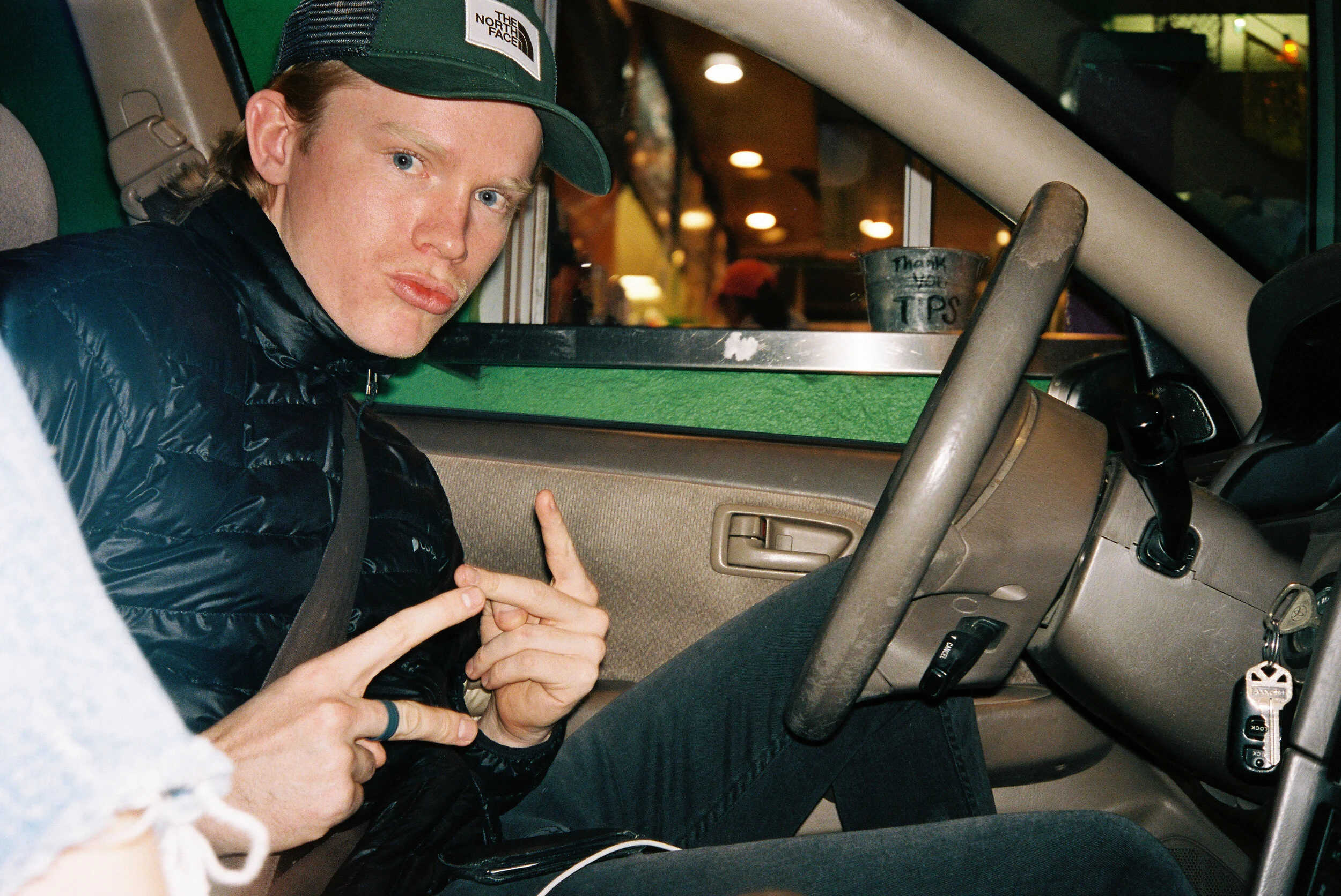 A young man with long blonde hair wearing a green baseball cap and black jacket sitting in a car at night, looking directly at the camera with a serious expression, inside a vehicle with a dashboard and steering wheel visible, and a metal tip jar lab