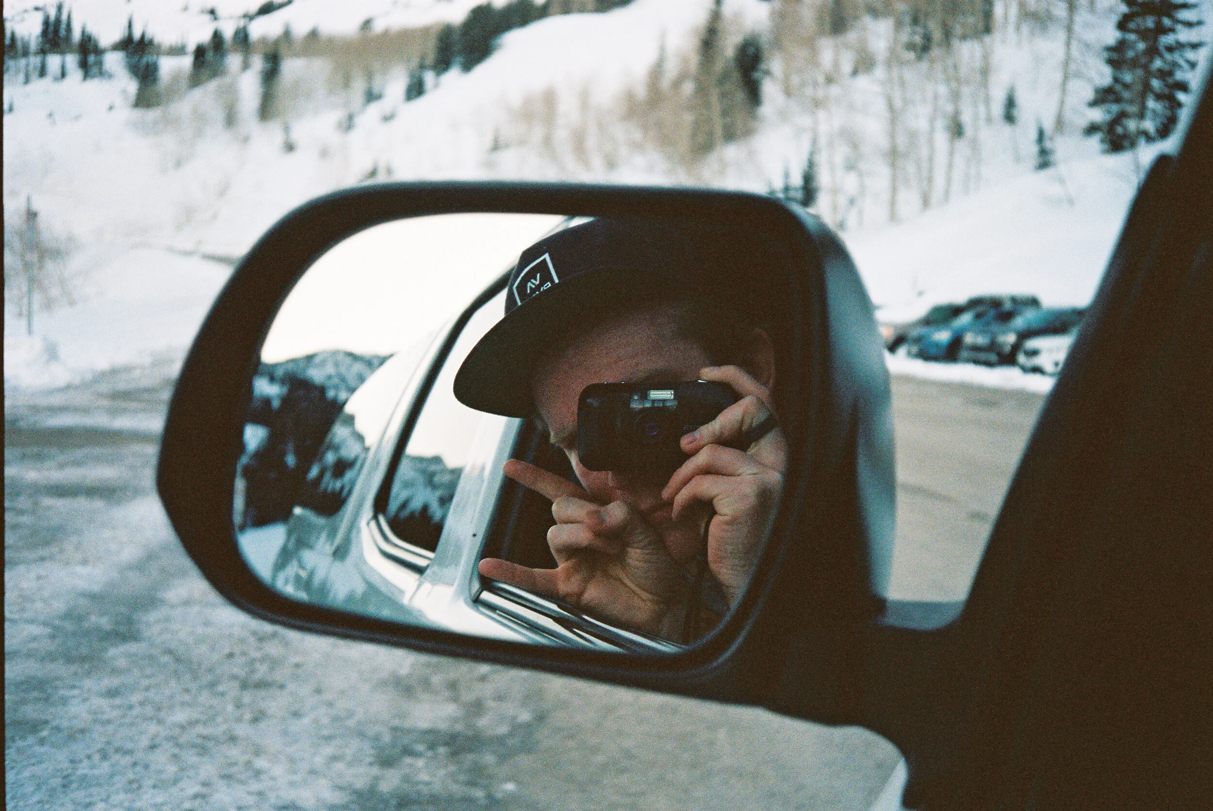 A person taking a selfie in a car's side mirror, wearing a black cap and holding a camera, with snow-covered trees and parked cars in the background.