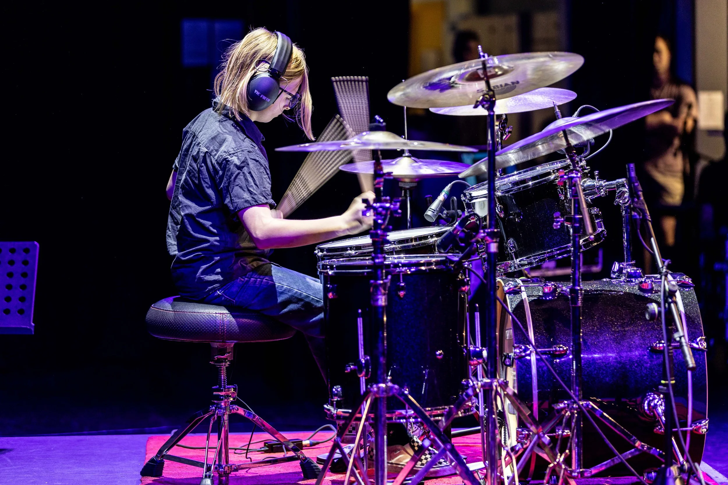 Young girl playing drums on stage with headphones, surrounded by cymbals and drum set, under purple stage lighting.