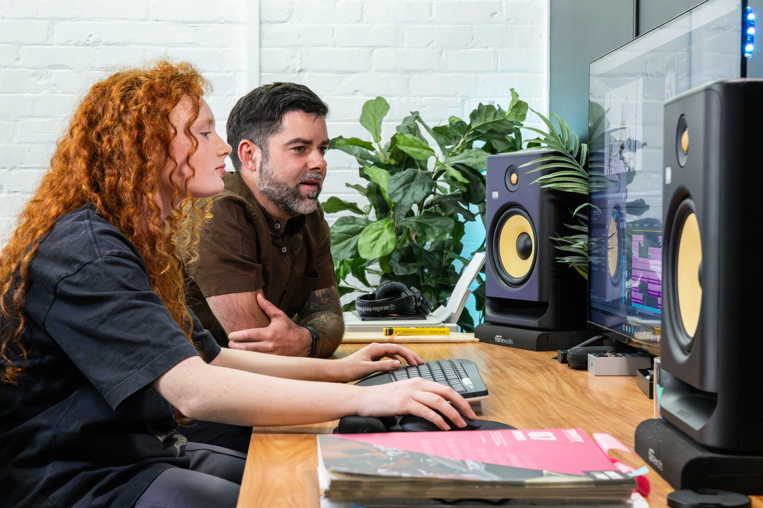 Two people working at a computer in a studio with speakers and plants in the background, one woman with curly red hair and a man with dark hair and a beard.