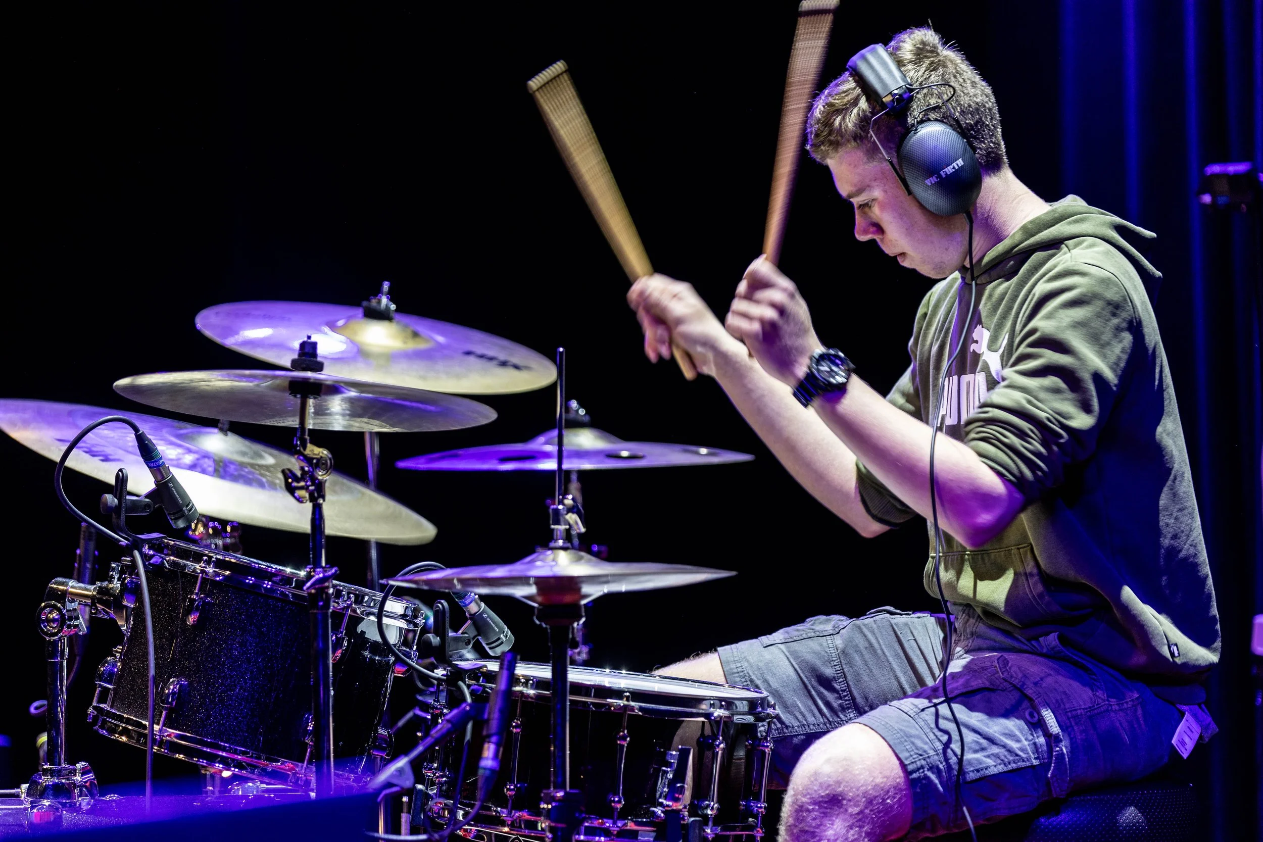 Young man playing drums with headphones on, sitting on a stool on stage, illuminated by purple stage lights.