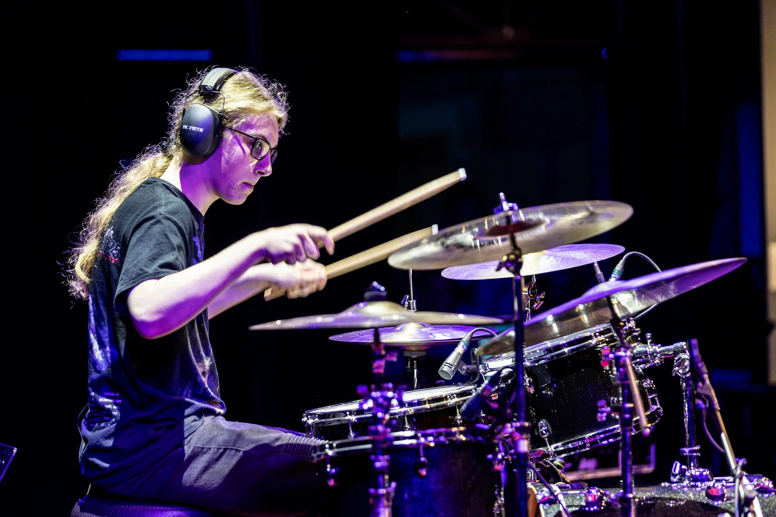 A young woman wearing headphones and glasses playing drum set on stage.