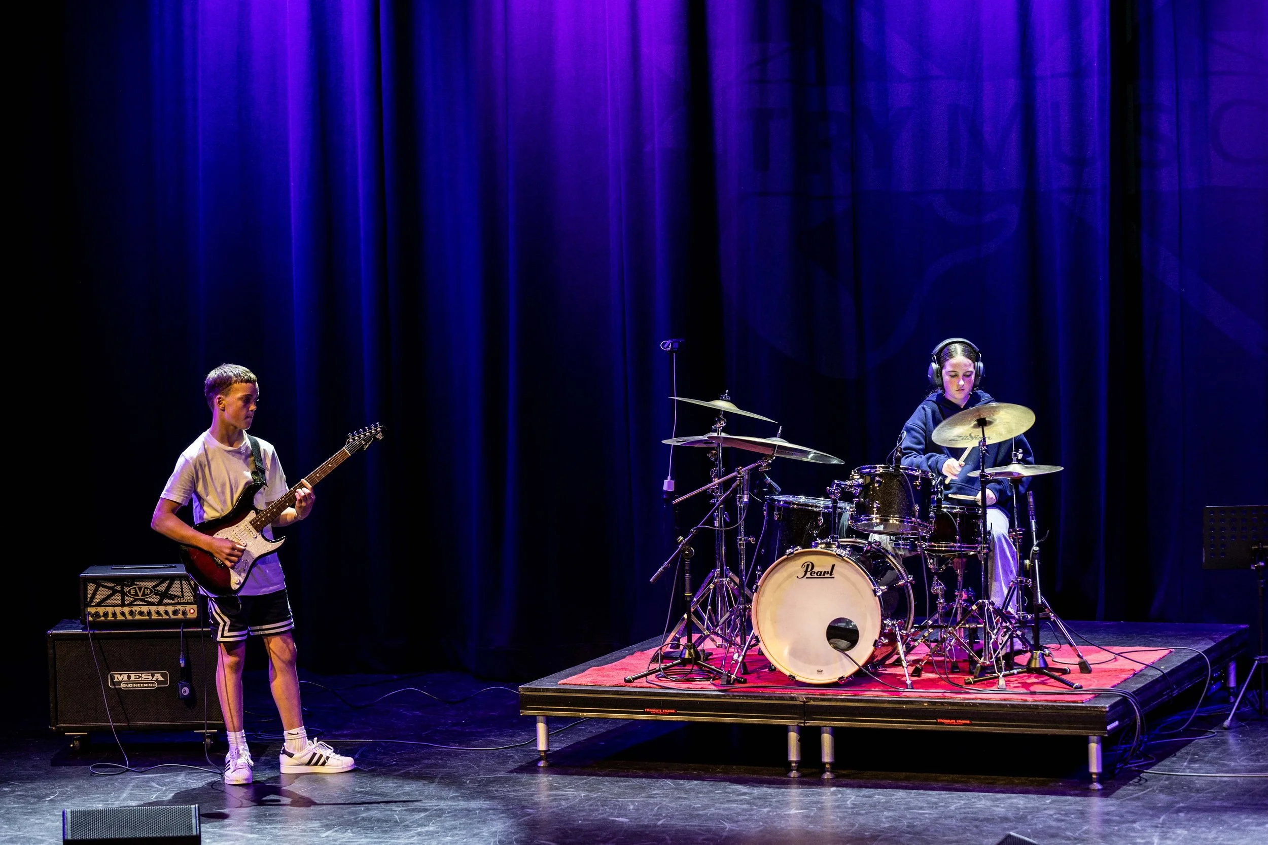 A young boy playing electric guitar and a girl playing drums on stage with a dark blue curtain backdrop