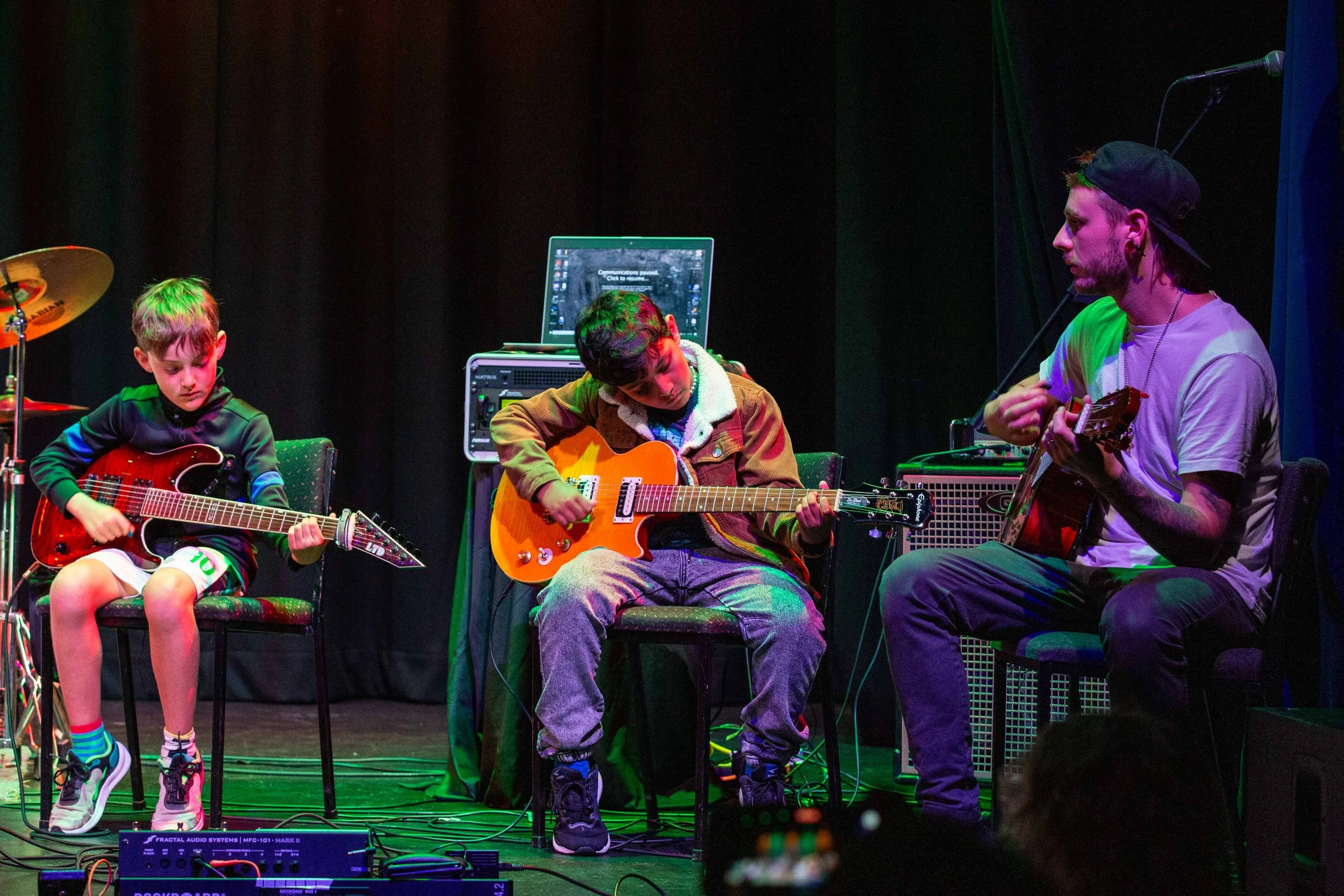 Three people, two children and an adult, playing guitars on stage during a performance, with musical equipment and a laptop behind them.