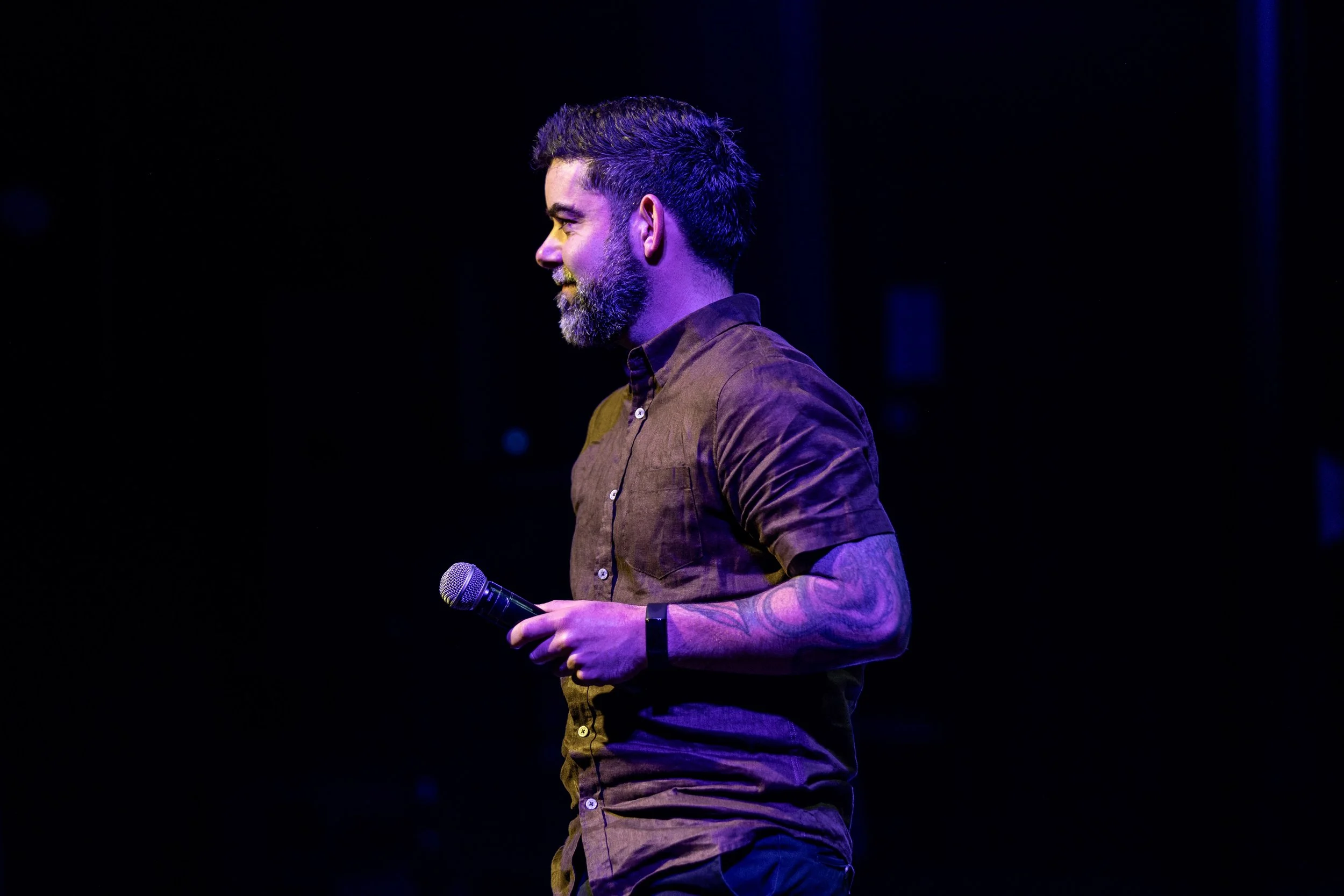 A man with a beard and short hair wearing a brown shirt holds a microphone on stage with dark background lighting.