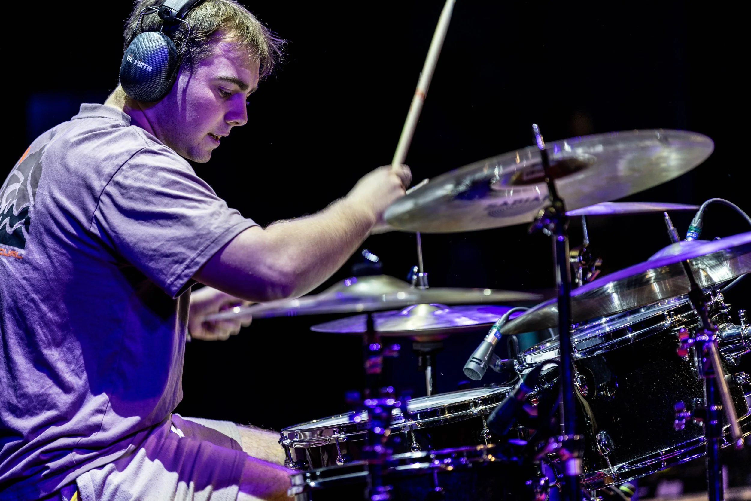 A male drummer wearing headphones playing drums on stage with dark background.