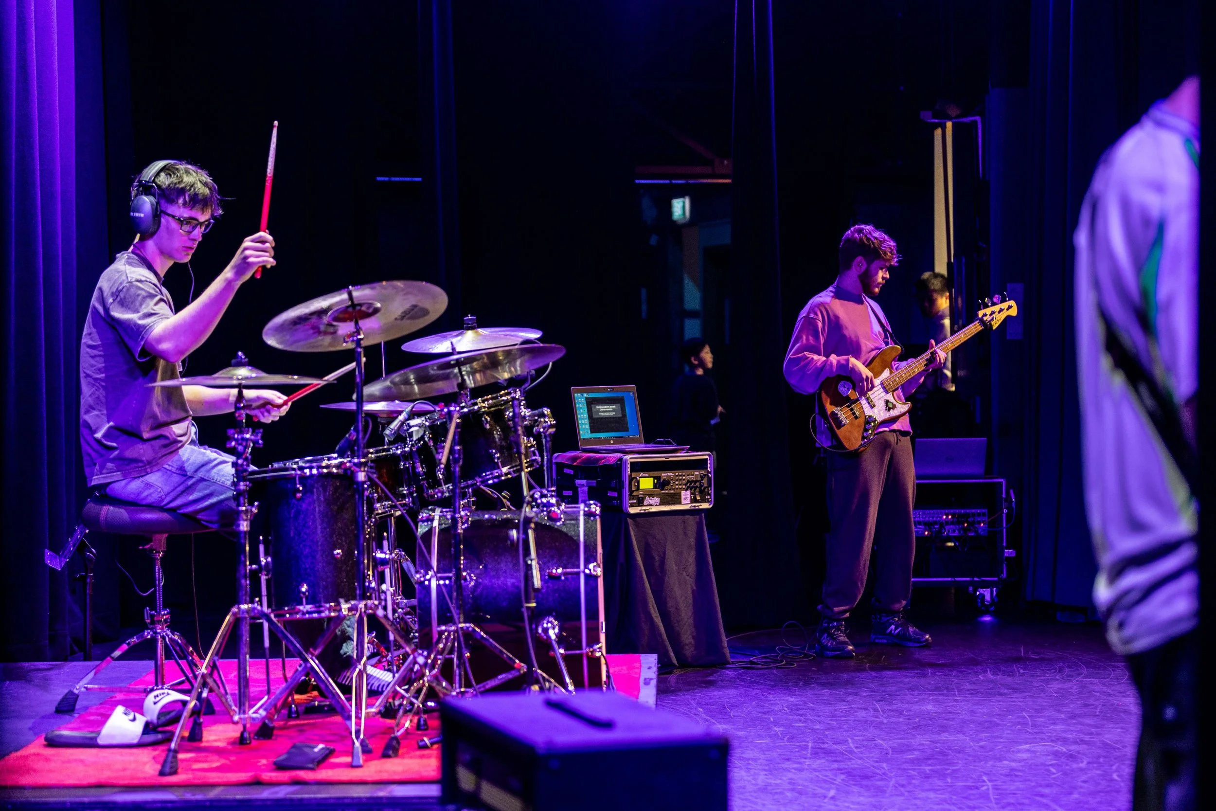 Music band rehearsing on stage with drummer and bassist, purple stage lighting.