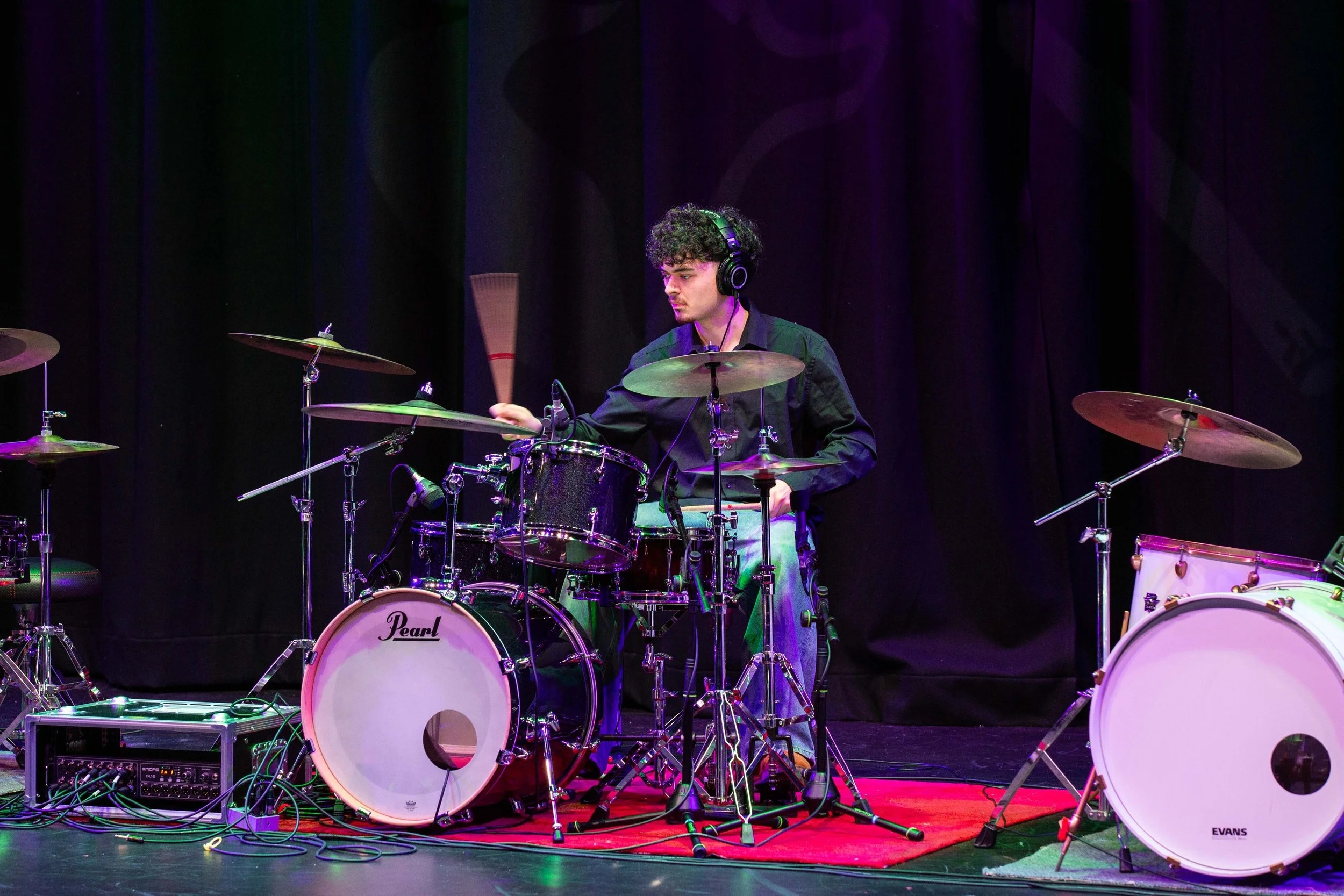 A young male drummer with curly hair wearing a black shirt and headphones playing on a stage with a black backdrop, using a Pearl drum kit with a red rug underneath, and with musical equipment and wires around.