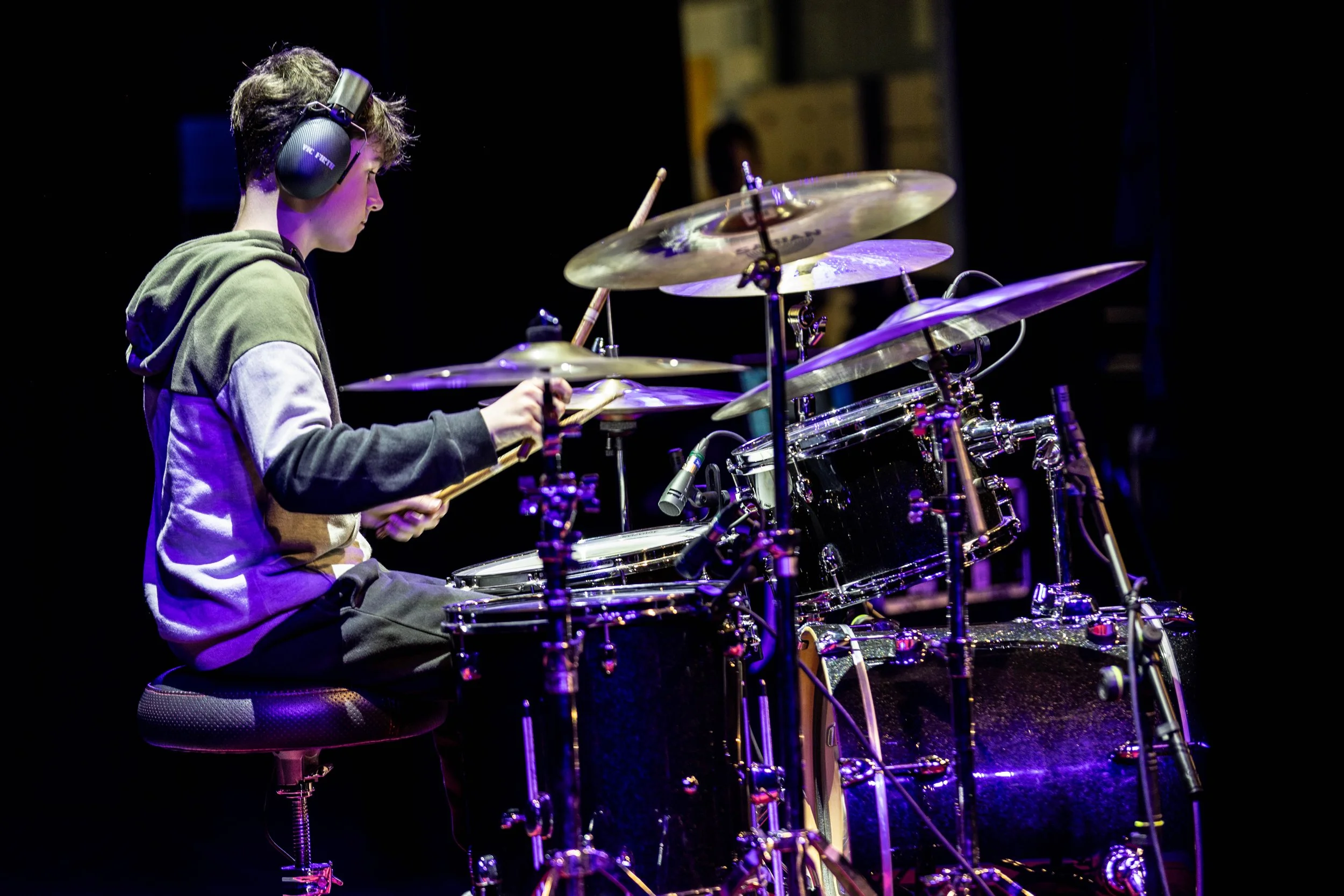 A young person with headphones playing a drum set on stage with purple lighting.