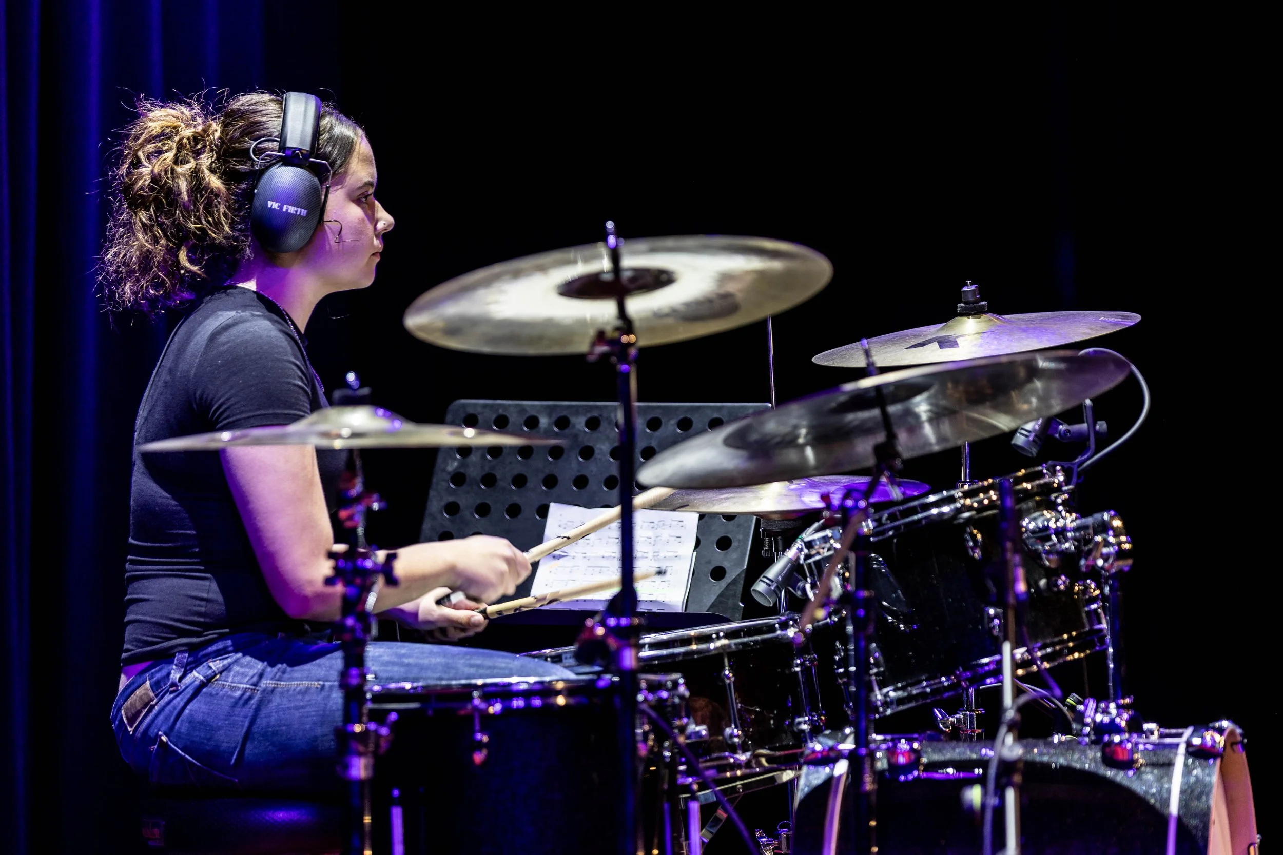 A woman playing drums on stage, wearing headphones, black shirt, and jeans, with sheet music on a stand in front of her.