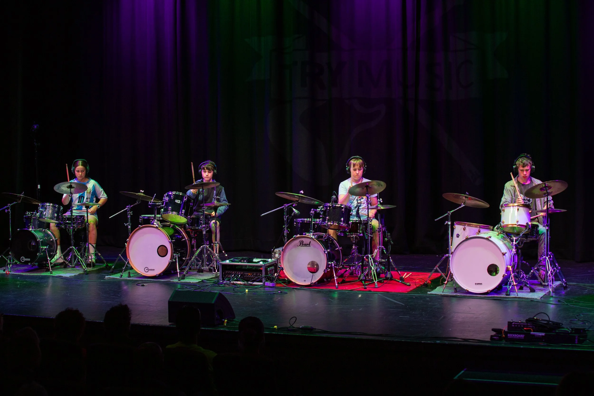 Four children playing drums on a stage with a dark curtain backdrop, colored stage lights, and an audience in foreground.
