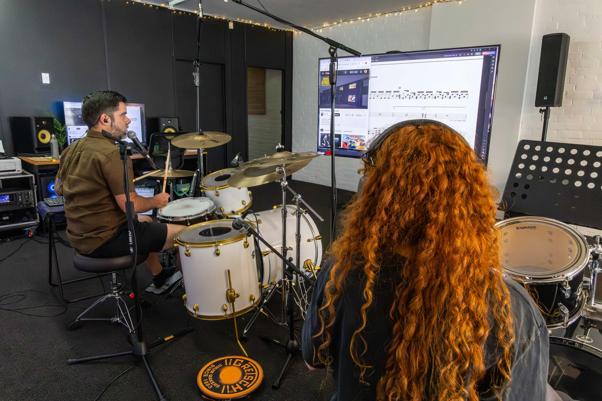 A music recording studio with a man sitting at a white drum set wearing headphones, and a woman with long, curly red hair wearing headphones. They are both facing a large screen displaying sheet music. The studio has black walls, music equipment, mon