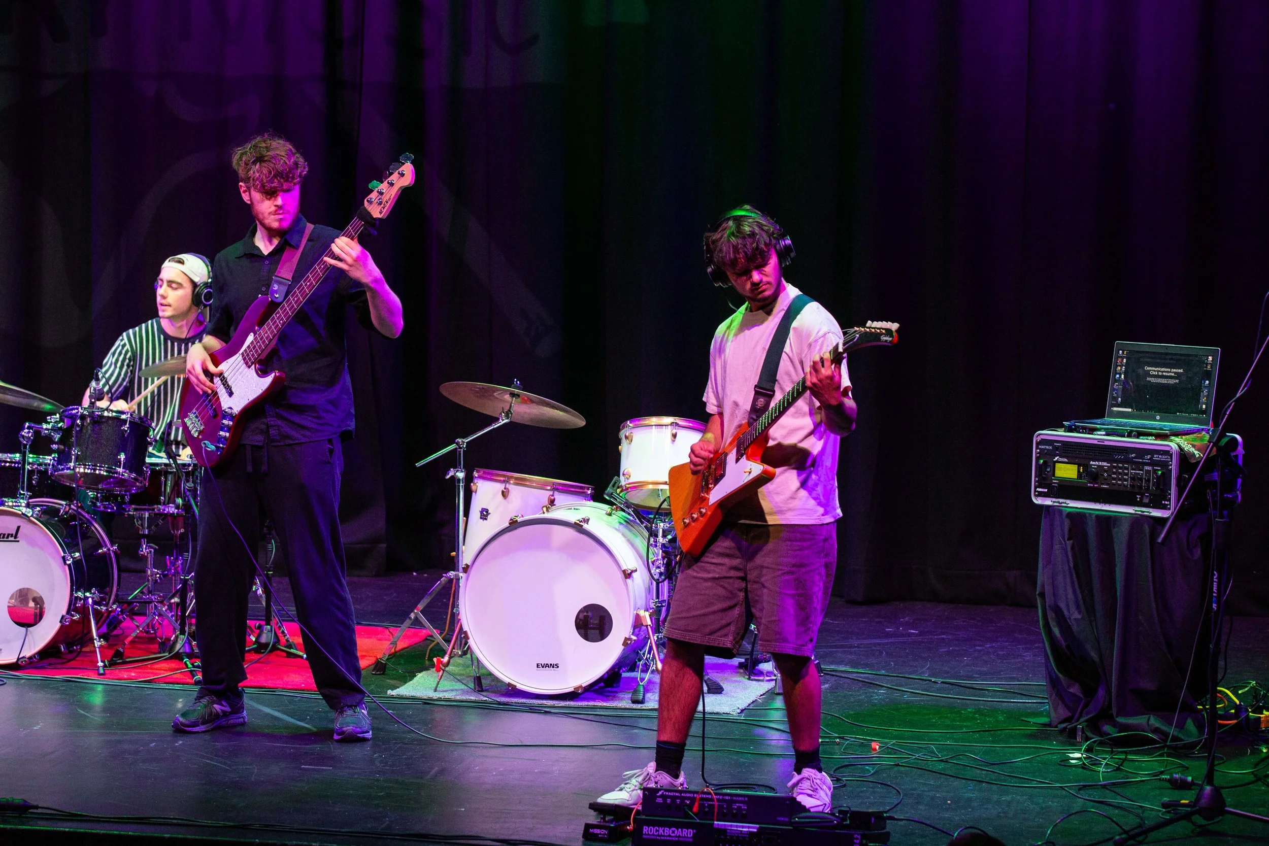 A band performs on a stage with purple lighting. The group includes a drummer, a bassist, and two guitarists, with equipment and a laptop on stage.