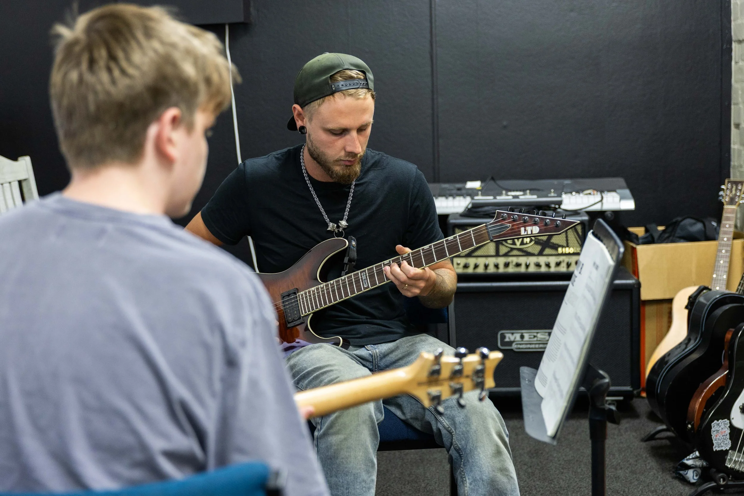 Two young men practicing guitar in a music room with guitar amplifiers and other guitars in the background.