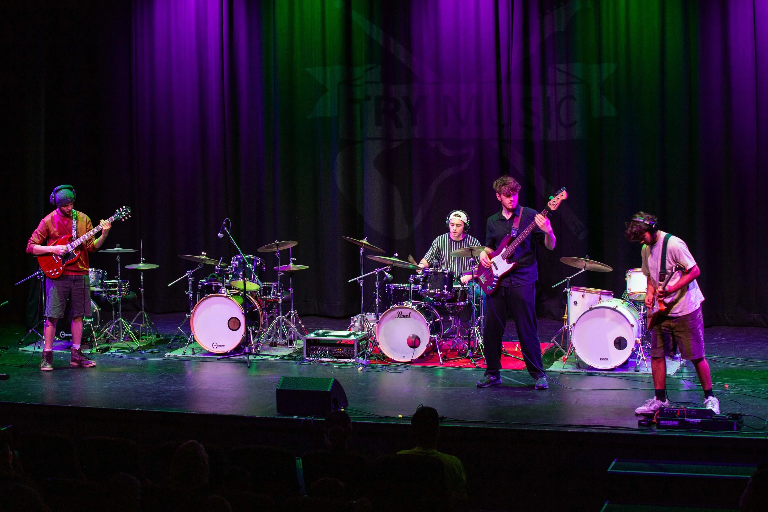 A band of five young musicians performs on stage, with colorful purple, green, and red lighting, and a black curtain backdrop.