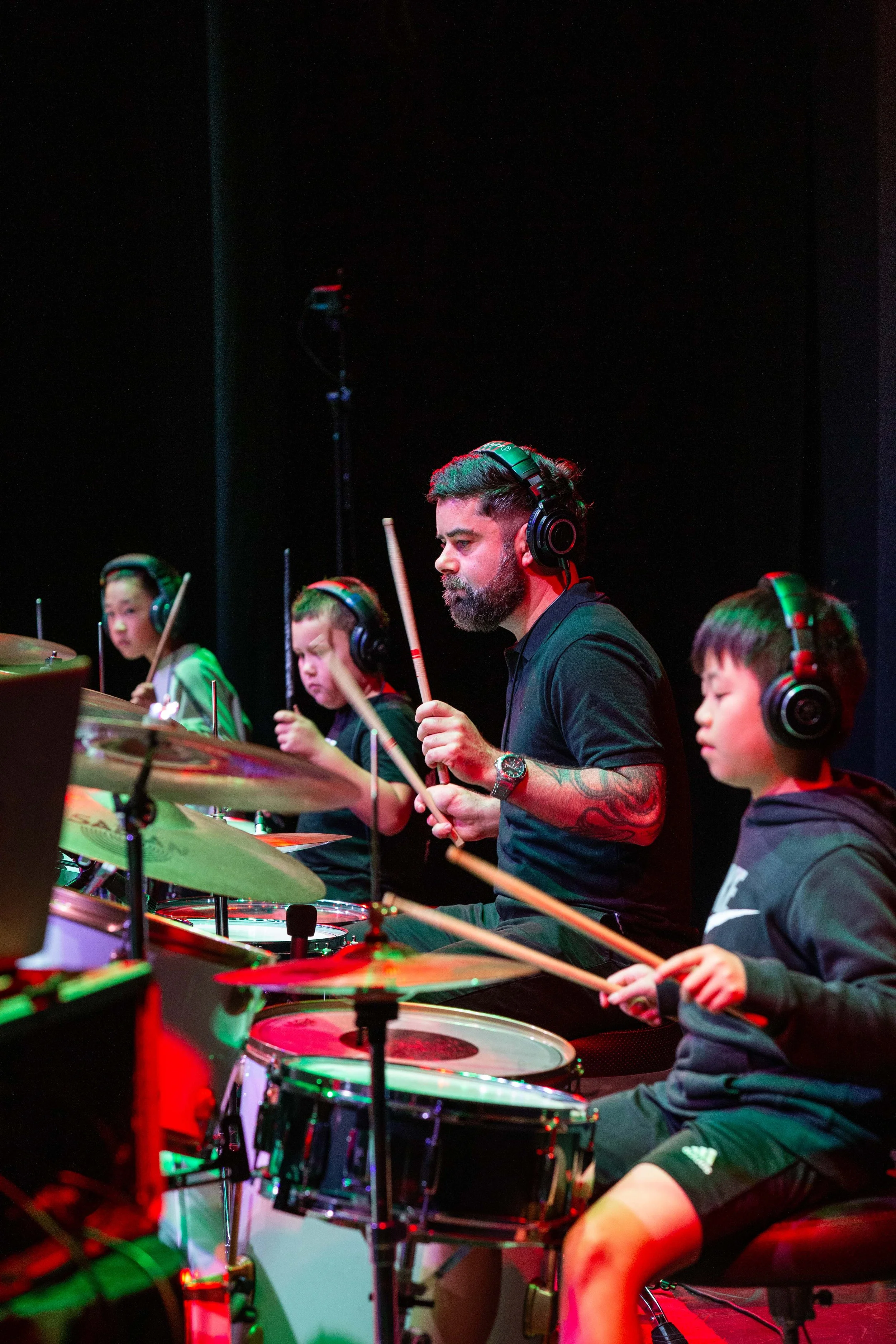 A music teacher with a beard and tattoos directs a group of children during a drum practice, all wearing headphones and holding drumsticks.