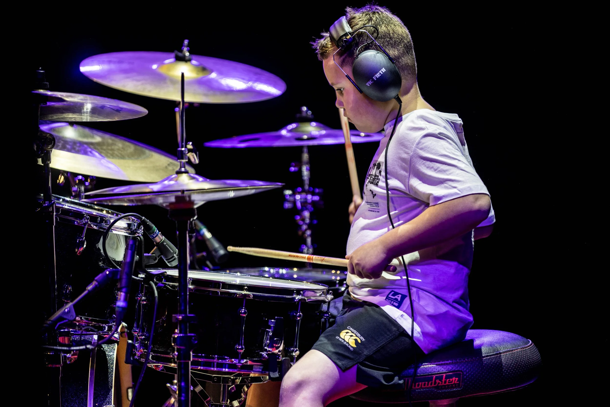 Young boy wearing headphones, playing drums on stage with purple lighting.