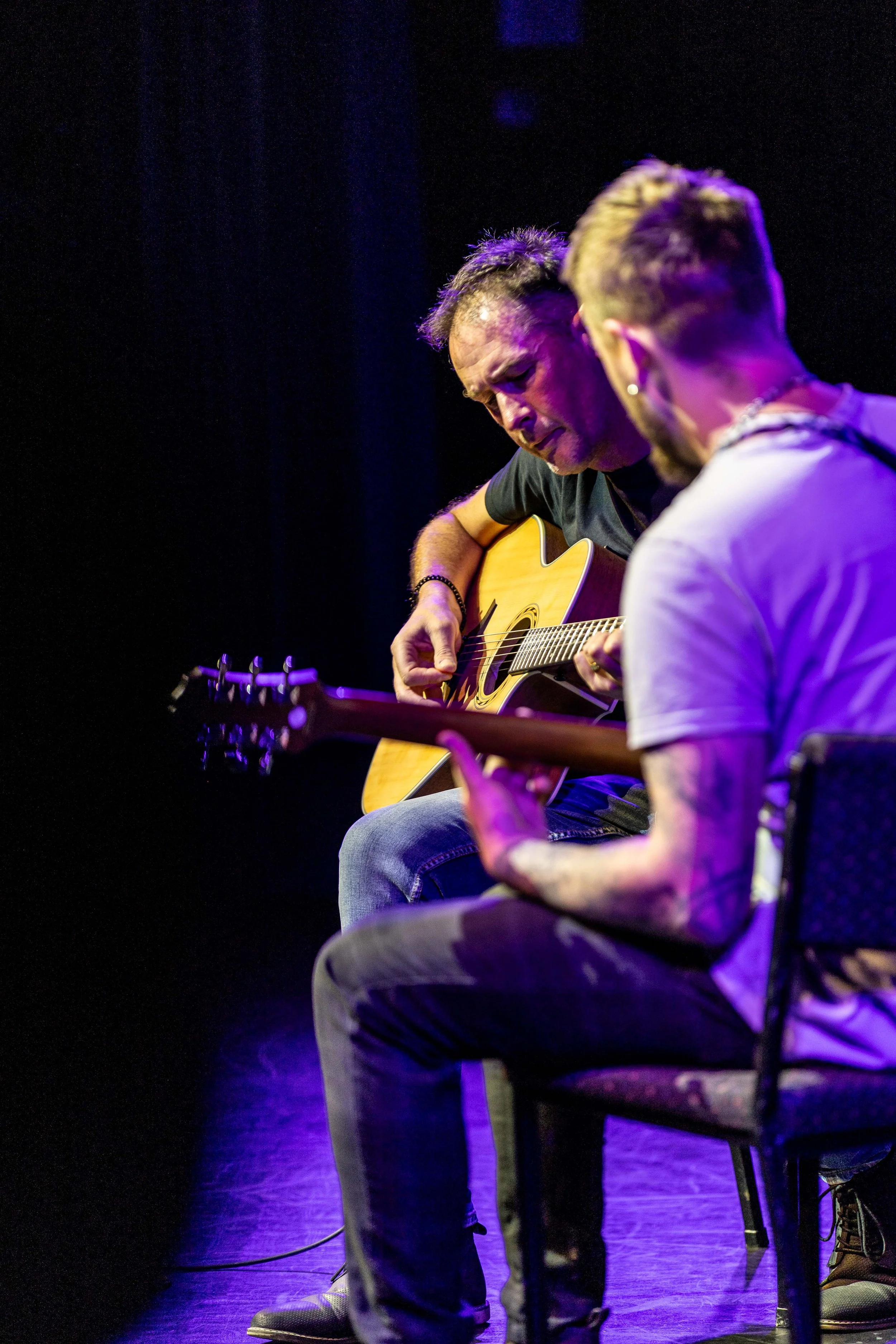 Two men playing acoustic guitars on stage in a dark setting with colorful lighting.