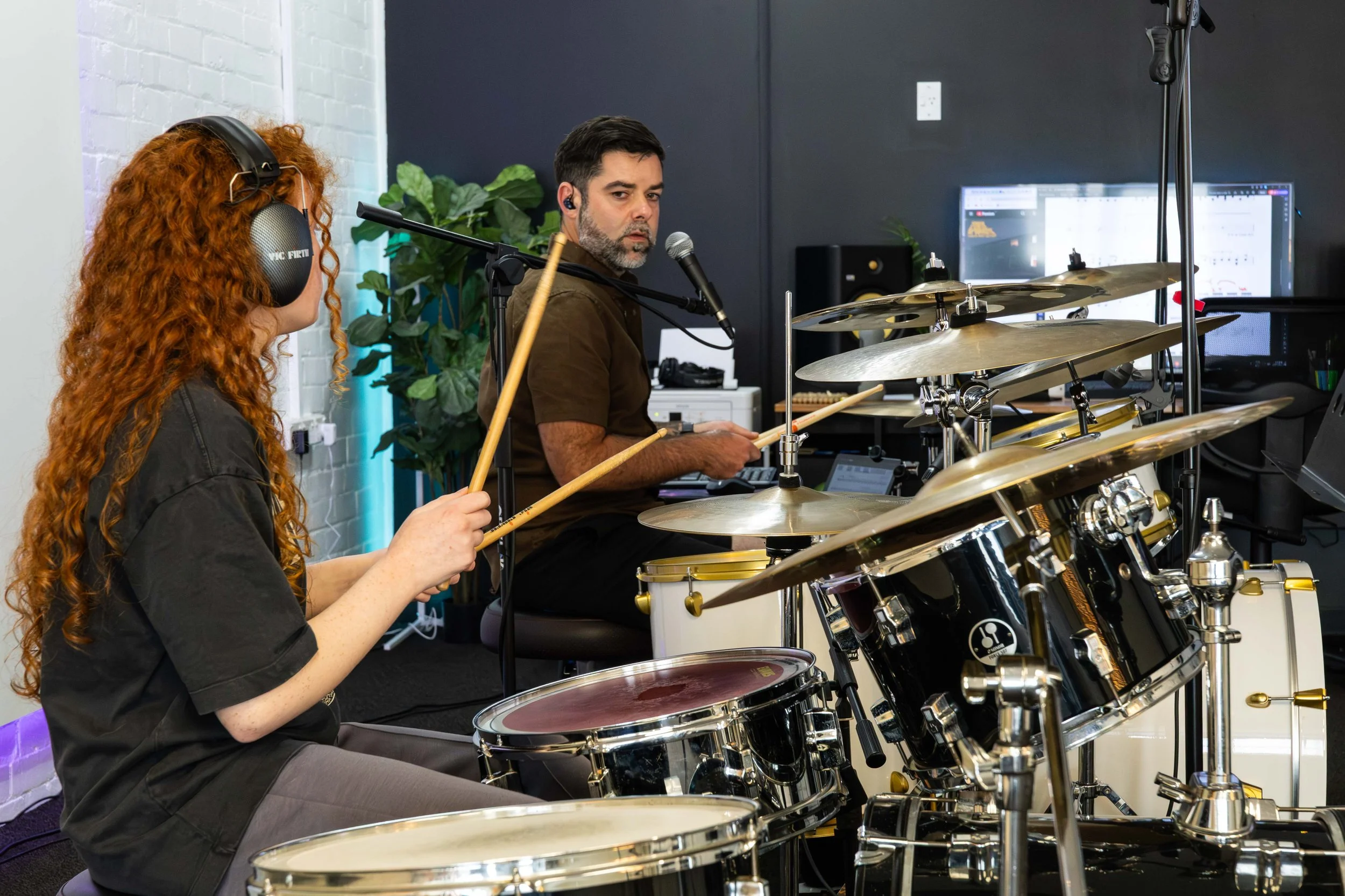 Two musicians, a woman with long curly red hair wearing headphones and a man with short dark hair and beard, playing drums in a music studio.