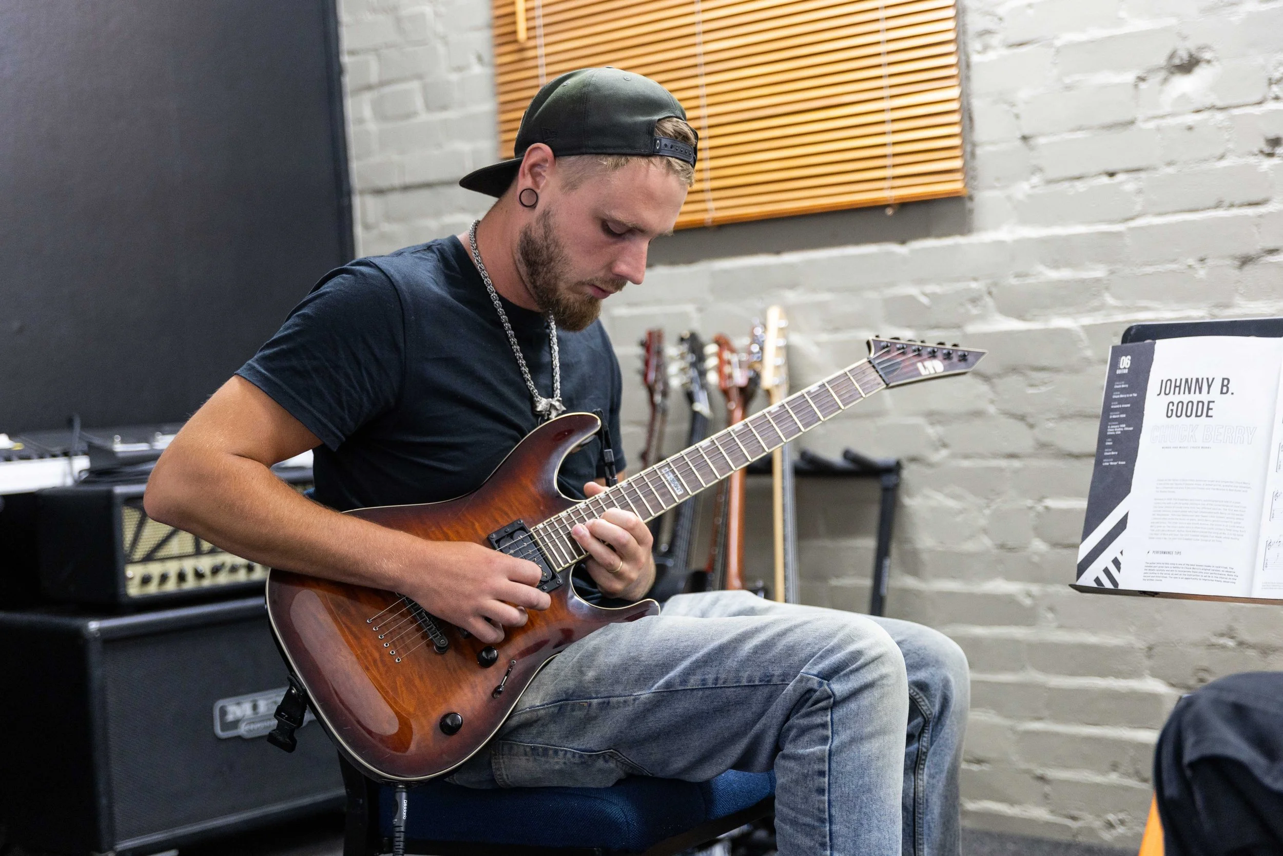 A man with a black cap, black t-shirt, and jeans playing an electric guitar in a music studio with guitars in the background.