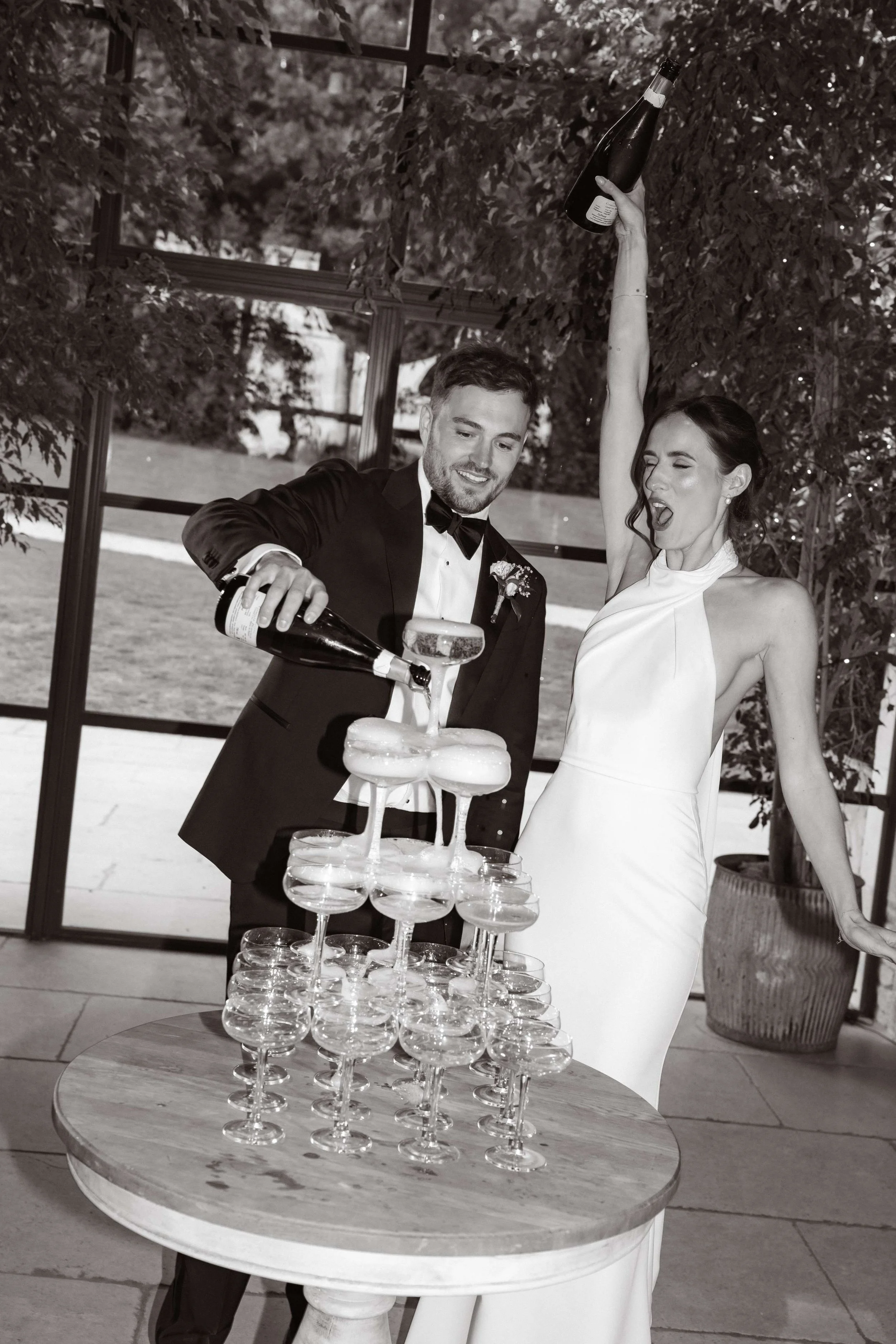 A bride and groom celebrating with a champagne tower at their wedding reception. The groom is pouring champagne into the top glass, and the bride is excited with one arm raised, holding a champagne bottle, in front of large windows showing trees outside.